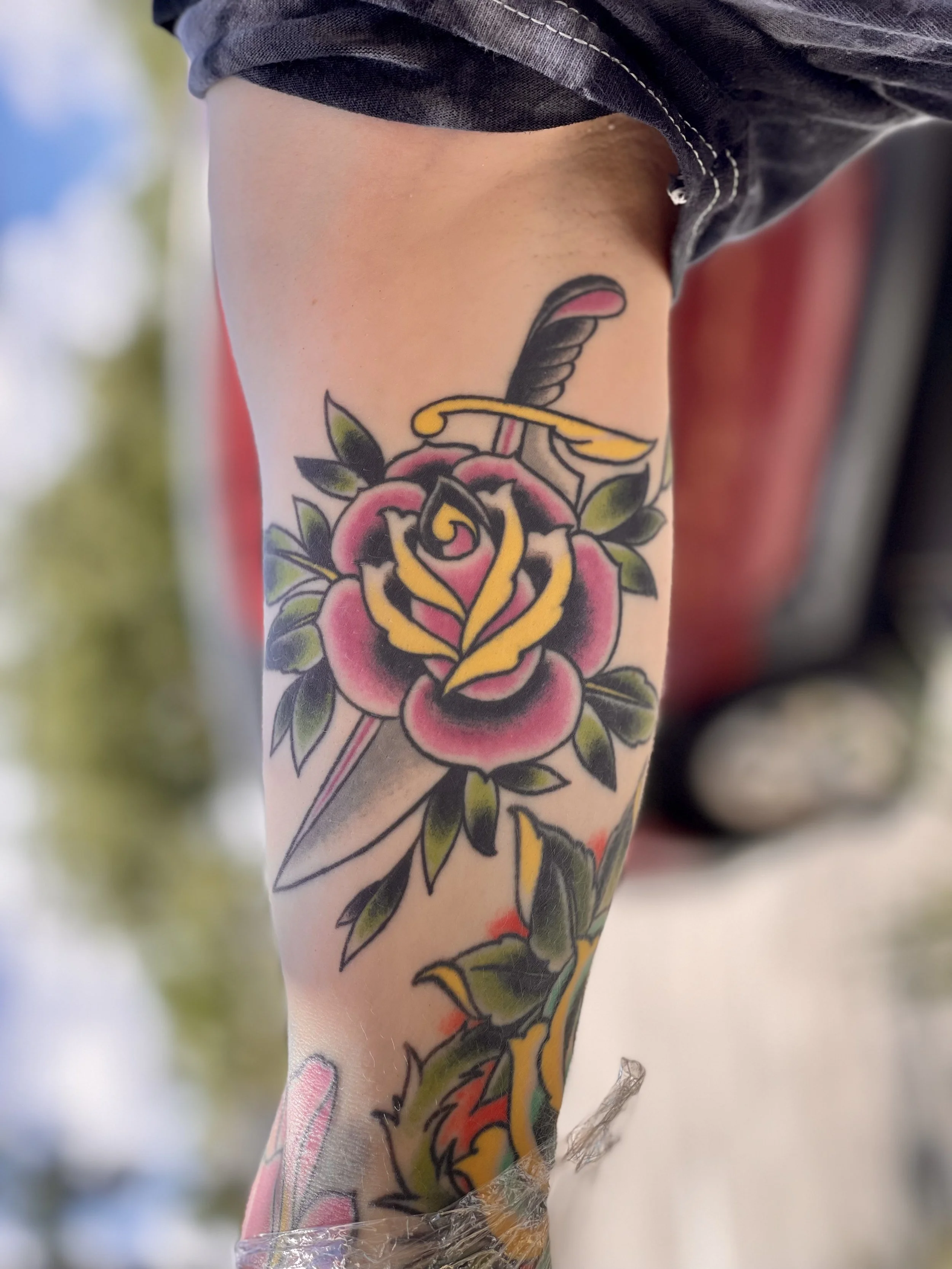 Healed classic traditional American color inner bicep tattoo featuring a bold pink and yellow rose pierced by a dagger with a gold crossguard and dark handle, surrounded by dark green leaves, custom tattoo by Nick at Big Kahuna Tattoo in Boca Raton, 