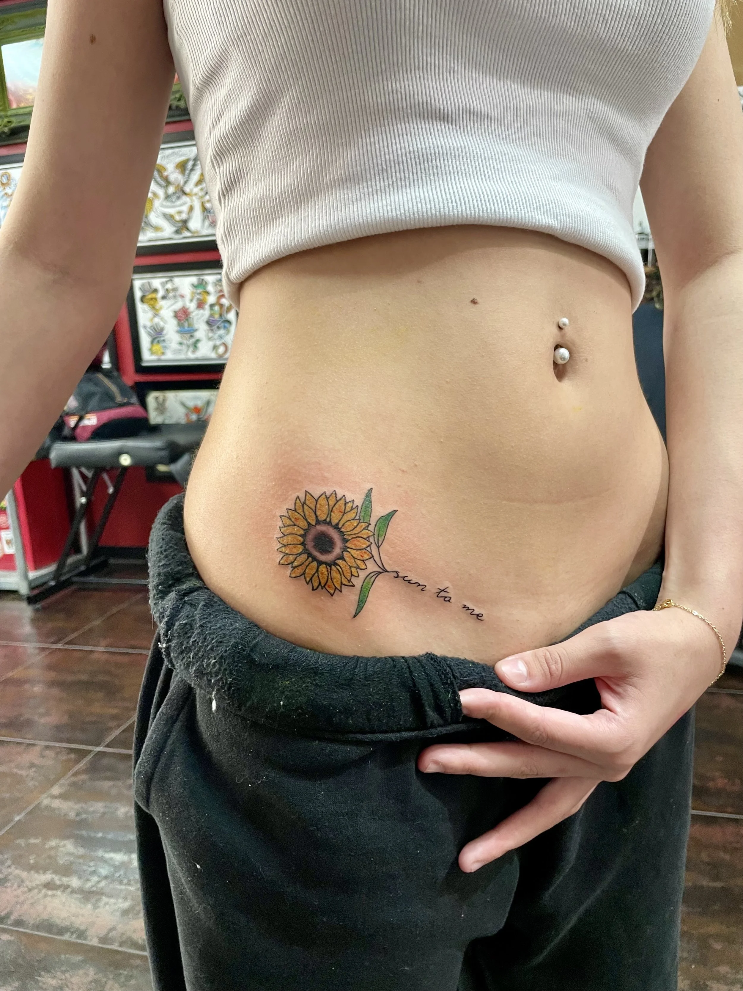 Fine line color hip tattoo featuring a sunflower with golden orange petals and a dark seeded center, with fine line script reading "sun to me" flowing along the stem, custom tattoo by Nick at Big Kahuna Tattoo in Boca Raton, FL.