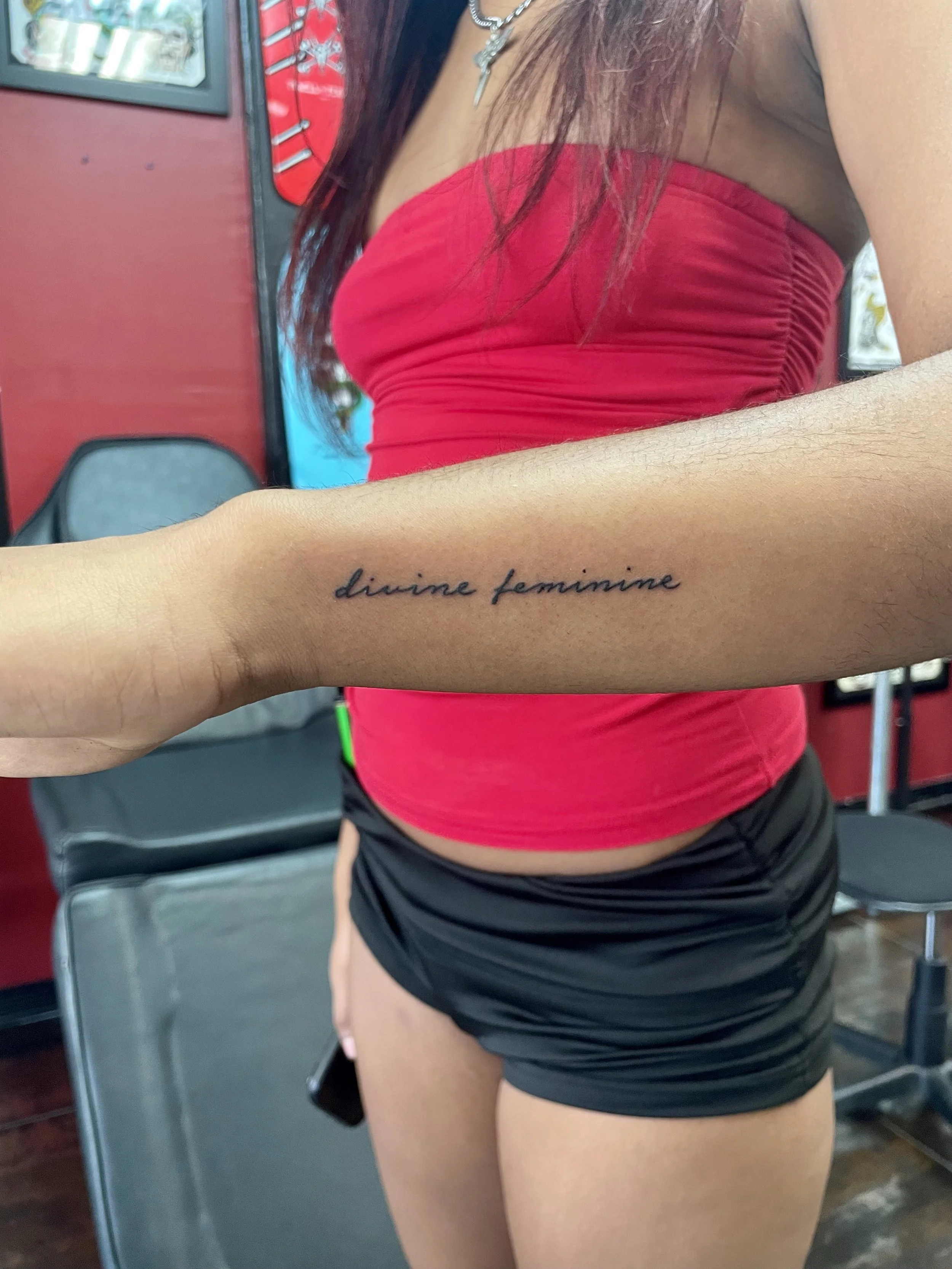 Fine line forearm tattoo featuring the phrase "divine feminine" in delicate flowing cursive lettering, custom tattoo by Nick at Big Kahuna Tattoo in Boca Raton, FL.