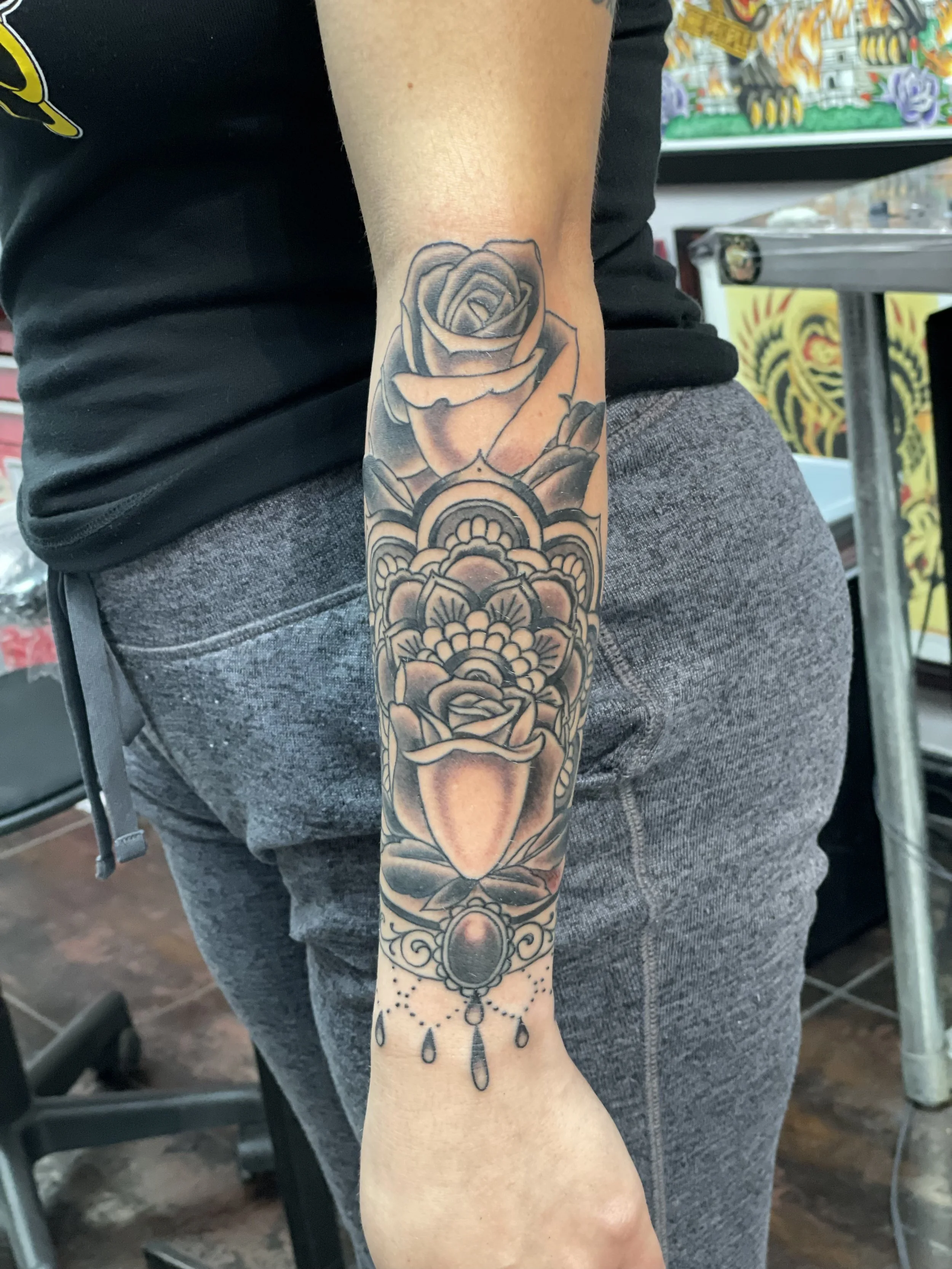 Black and grey forearm tattoo featuring a traditional rose at the elbow flowing into a detailed geometric mandala at the center, transitioning into a second rose at the wrist with an ornate jeweled pendant and drop chain detail below, custom tattoo b