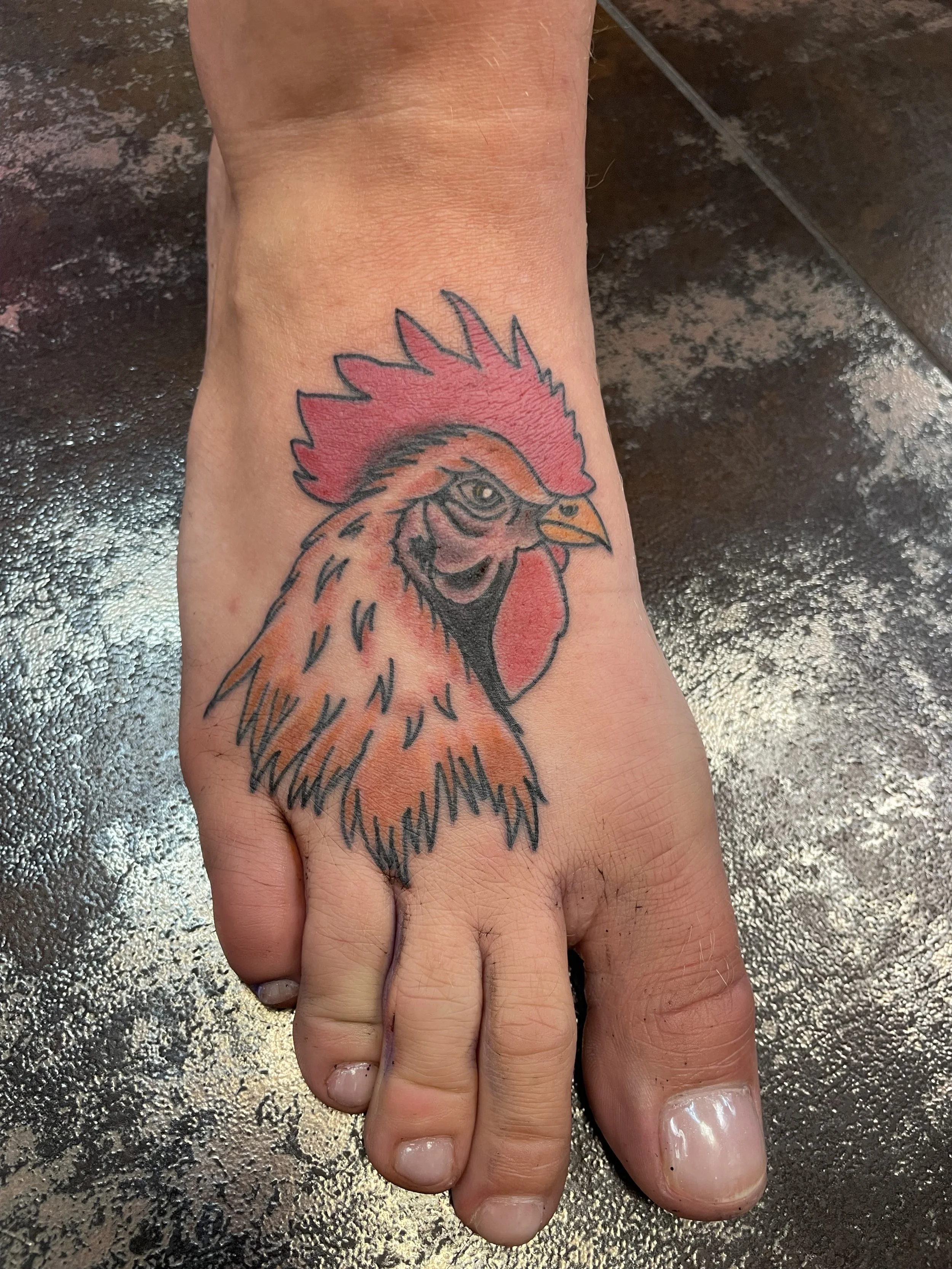 Traditional American color foot tattoo featuring a bold rooster head portrait with a vivid red comb, orange and brown feathers, and a yellow beak, custom tattoo by Nick at Big Kahuna Tattoo in Boca Raton, FL.
