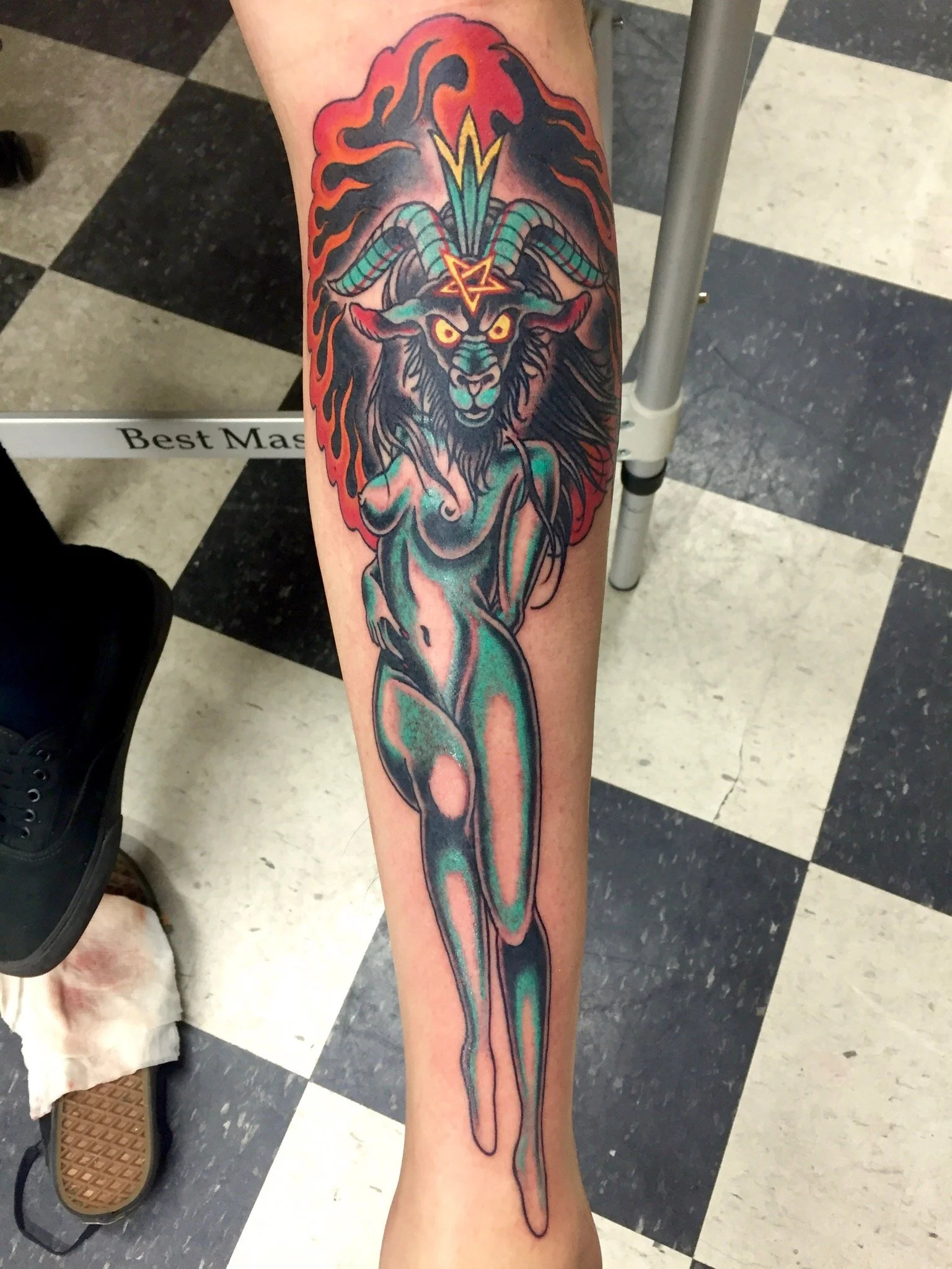 Traditional American color shin tattoo featuring a Baphomet head on the body of a pin up girl, rendered in green with a pentagram, ram horns, and a ring of red and orange flames surrounding the top, custom tattoo by Nick at Big Kahuna Tattoo in Boca 
