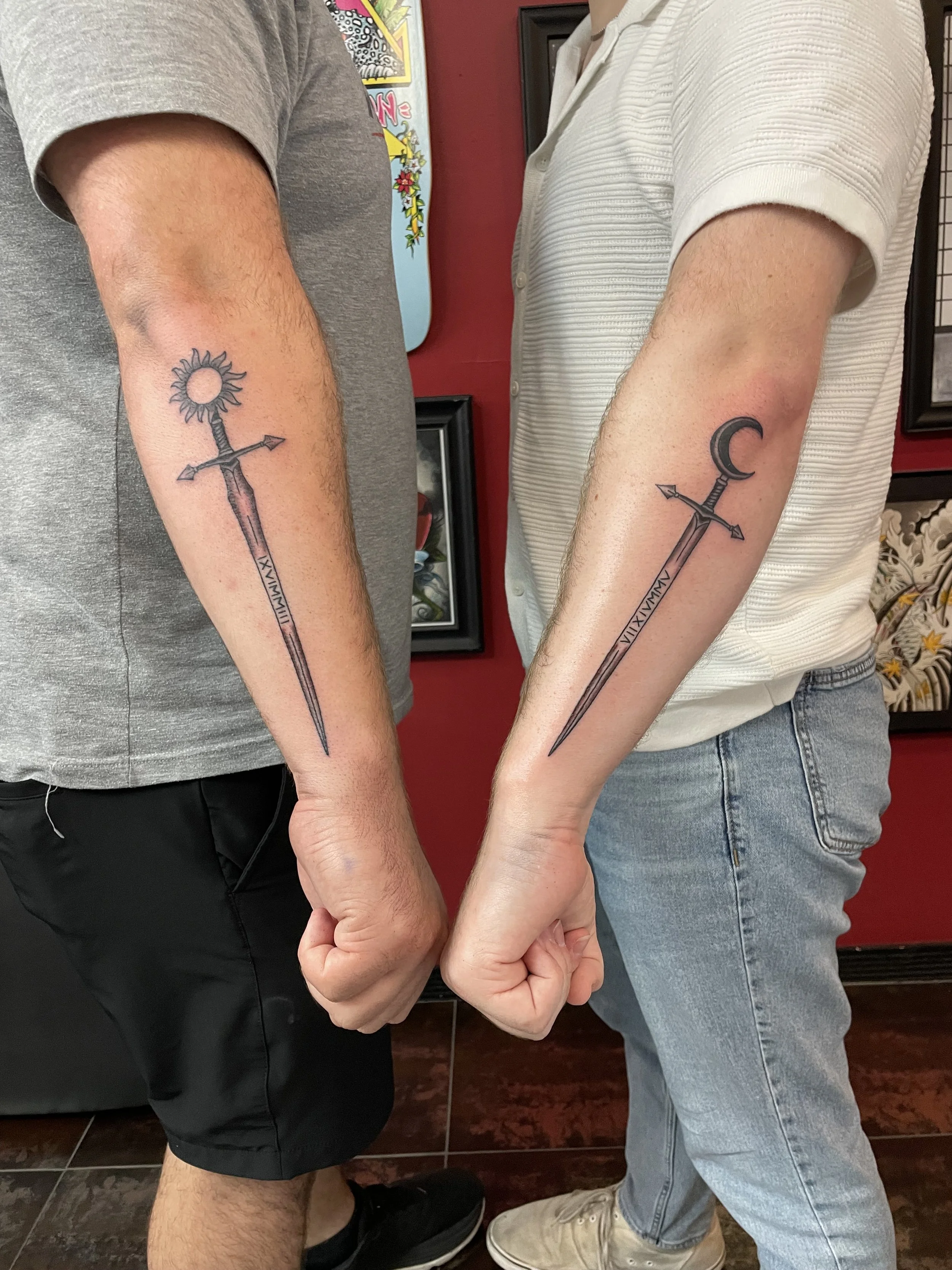 Matching black and grey forearm tattoos for two, featuring a pair of ornate daggers — one crowned with a radiant sun and one with a crescent moon — each with arrow-tipped crossguards and Roman numerals engraved along the blade, custom matching tattoo