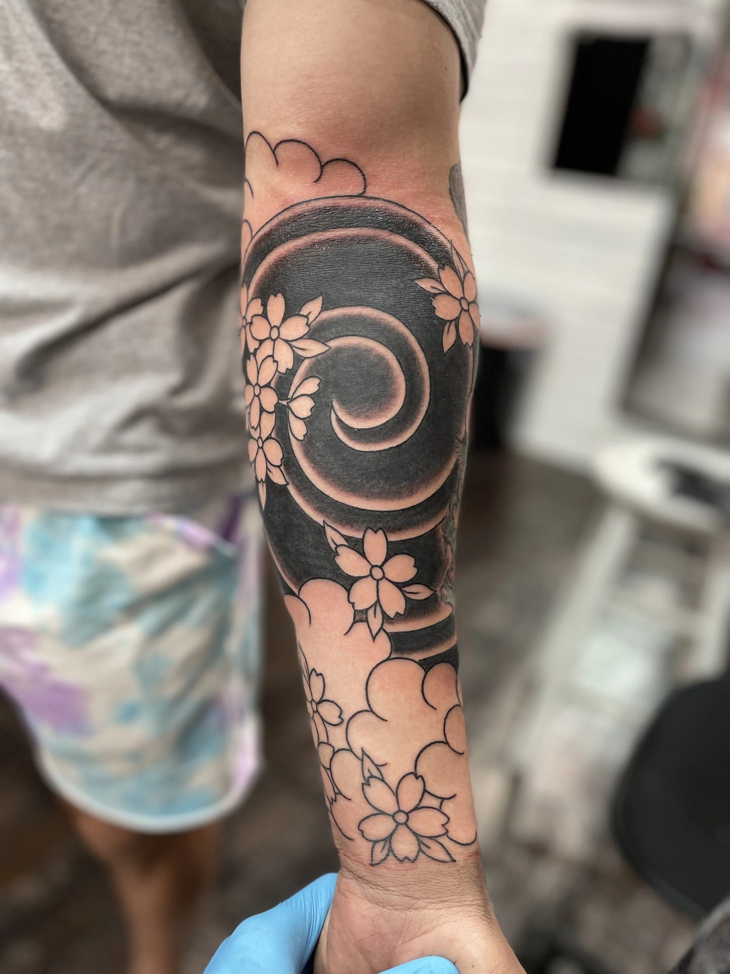 A bold spiraling Japanese cloud swirl in deep black occupies the center of the forearm, with delicate negative space cherry blossoms scattered throughout. A striking balance of heavy black and open skin. Japanese black and grey forearm tattoo by Nick