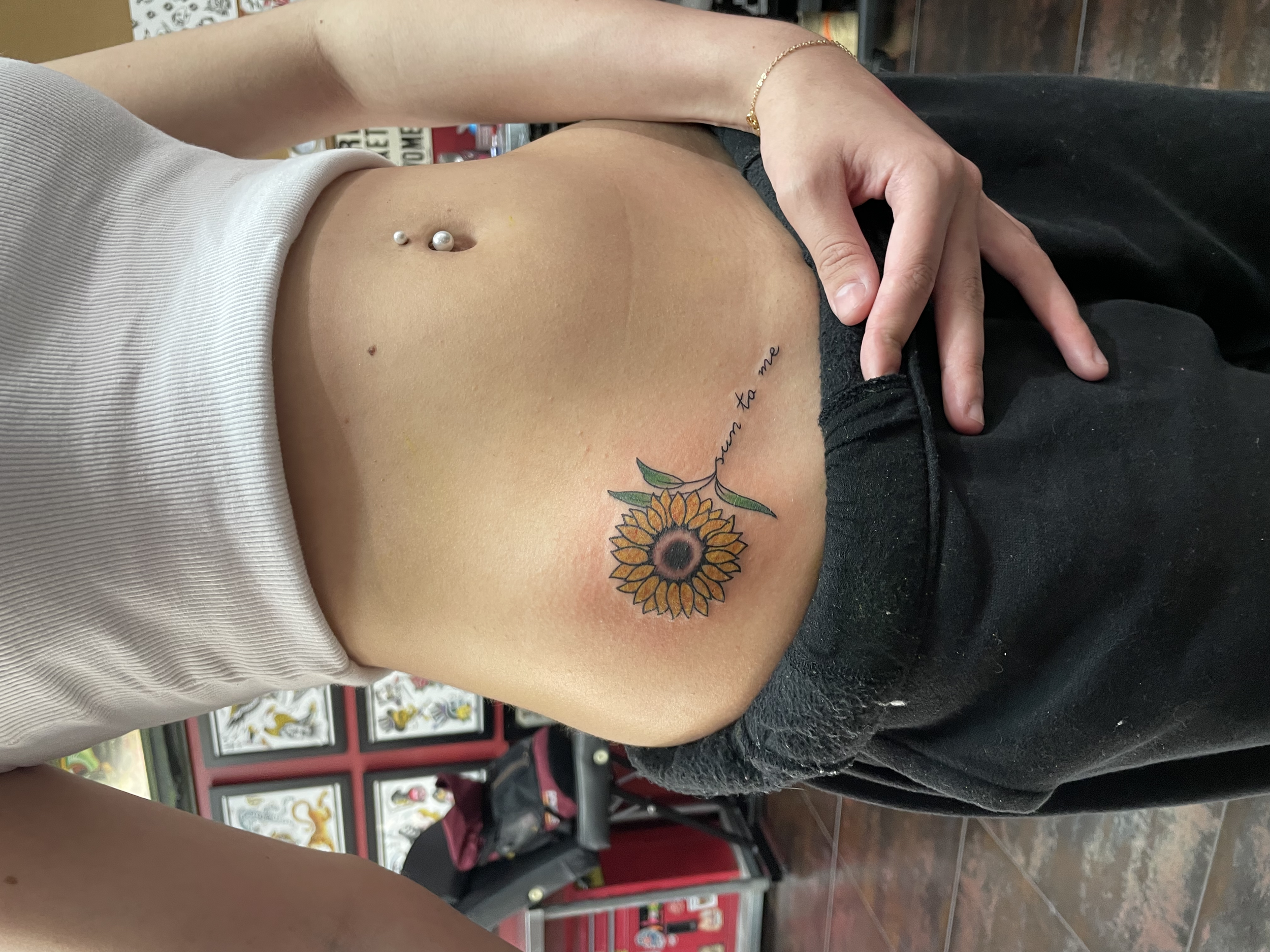 Color hip tattoo featuring a bright yellow sunflower with the cursive script "sun to me" flowing along the stem, custom tattoo by Nick at Big Kahuna Tattoo in Boca Raton, FL.