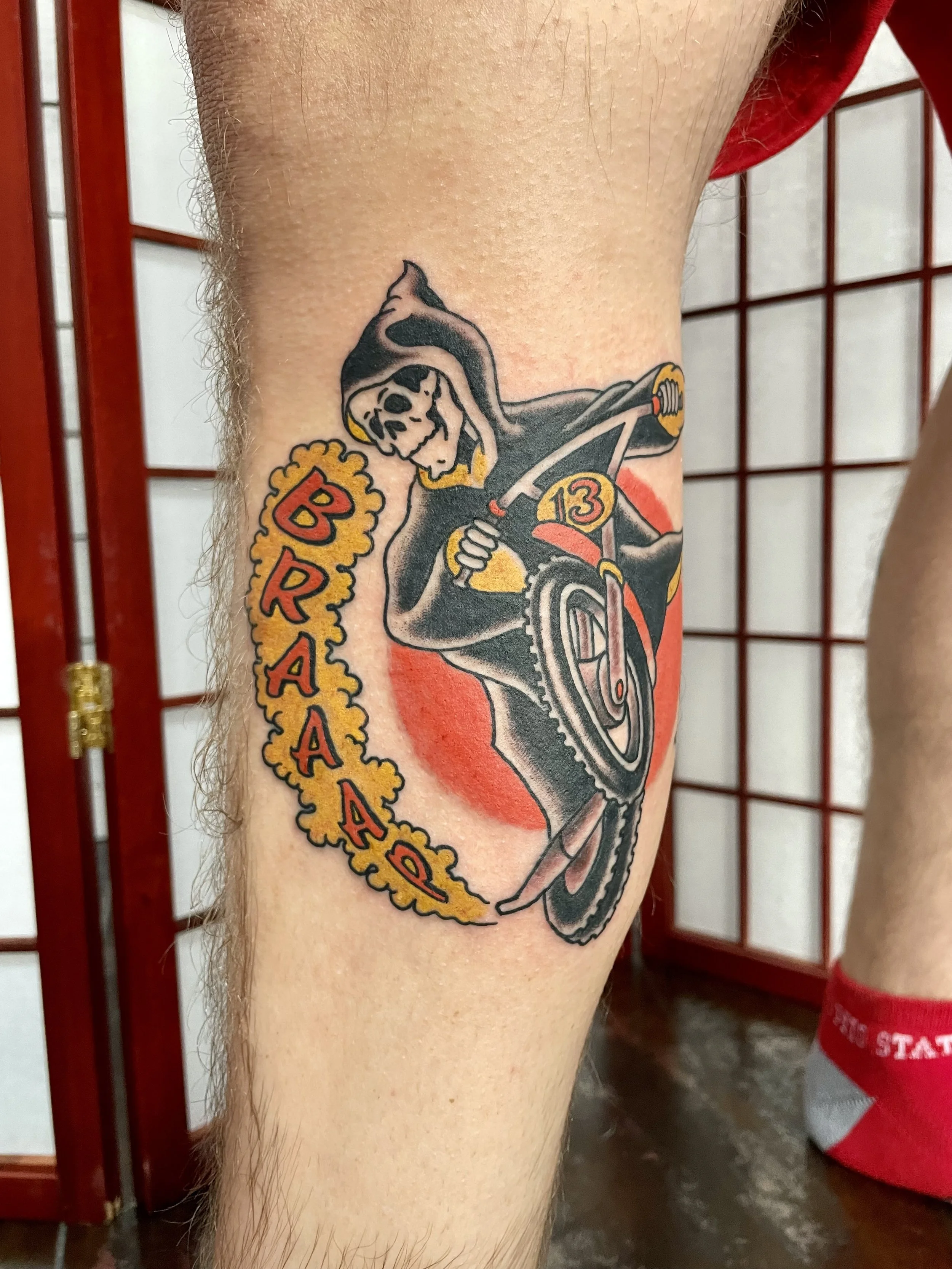 Death on a dirt bike! A hooded Grim Reaper leaning into a number 13 motorcycle surrounded by a gold exhaust smoke border spelling out "BRAAP." Fun, dark, and drawn to fit. Traditional American color leg tattoo by Nick at Big Kahuna Tattoo in Boca Rat
