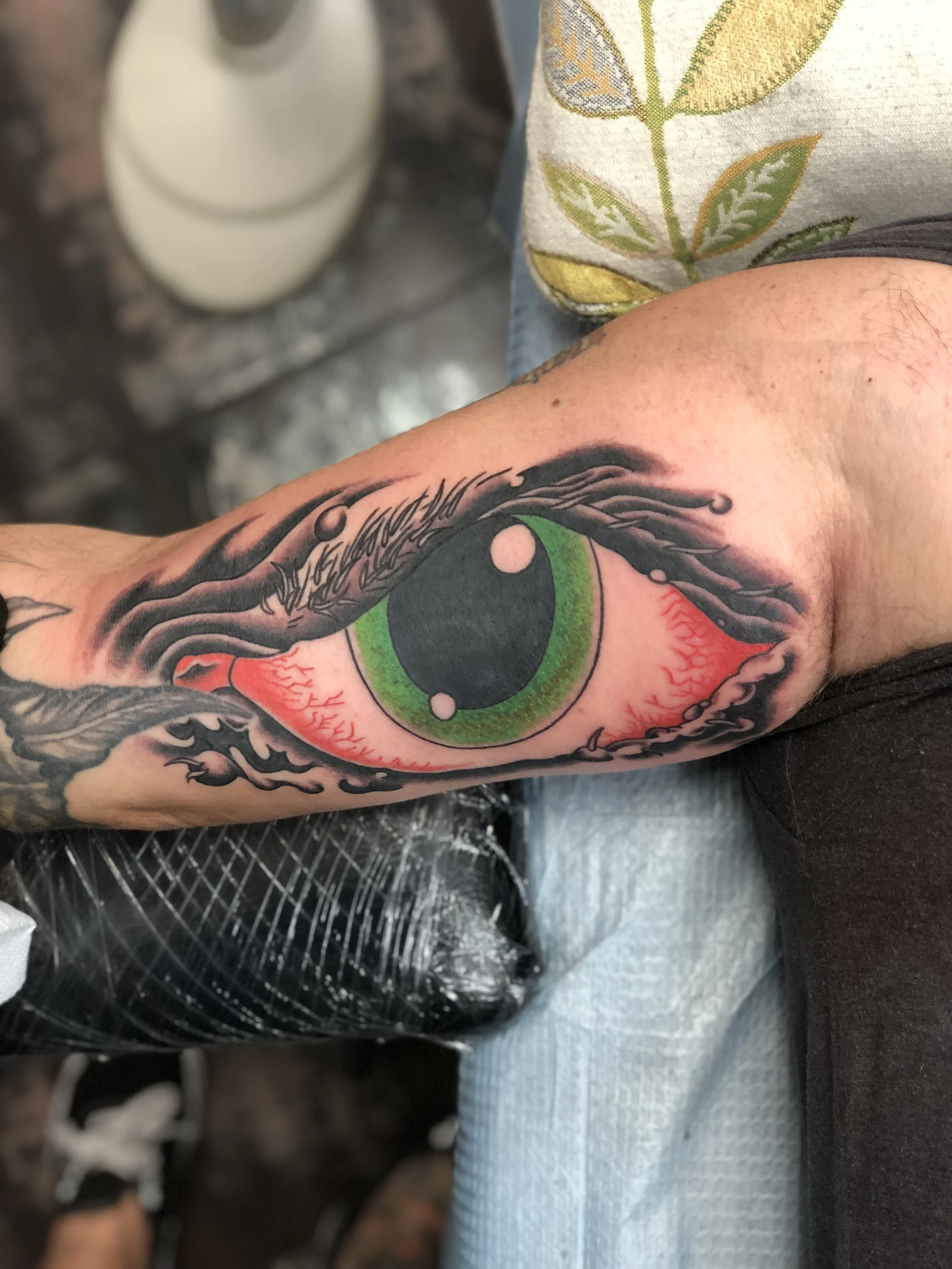 Traditional inner bicep tattoo featuring a tribute to the iconic Rob Roskopp eye graphic, originally illustrated by Jim Phillips for Santa Cruz Skateboards in the 1980s, with a bold green iris and bloodshot veins surrounded by dark dramatic linework,