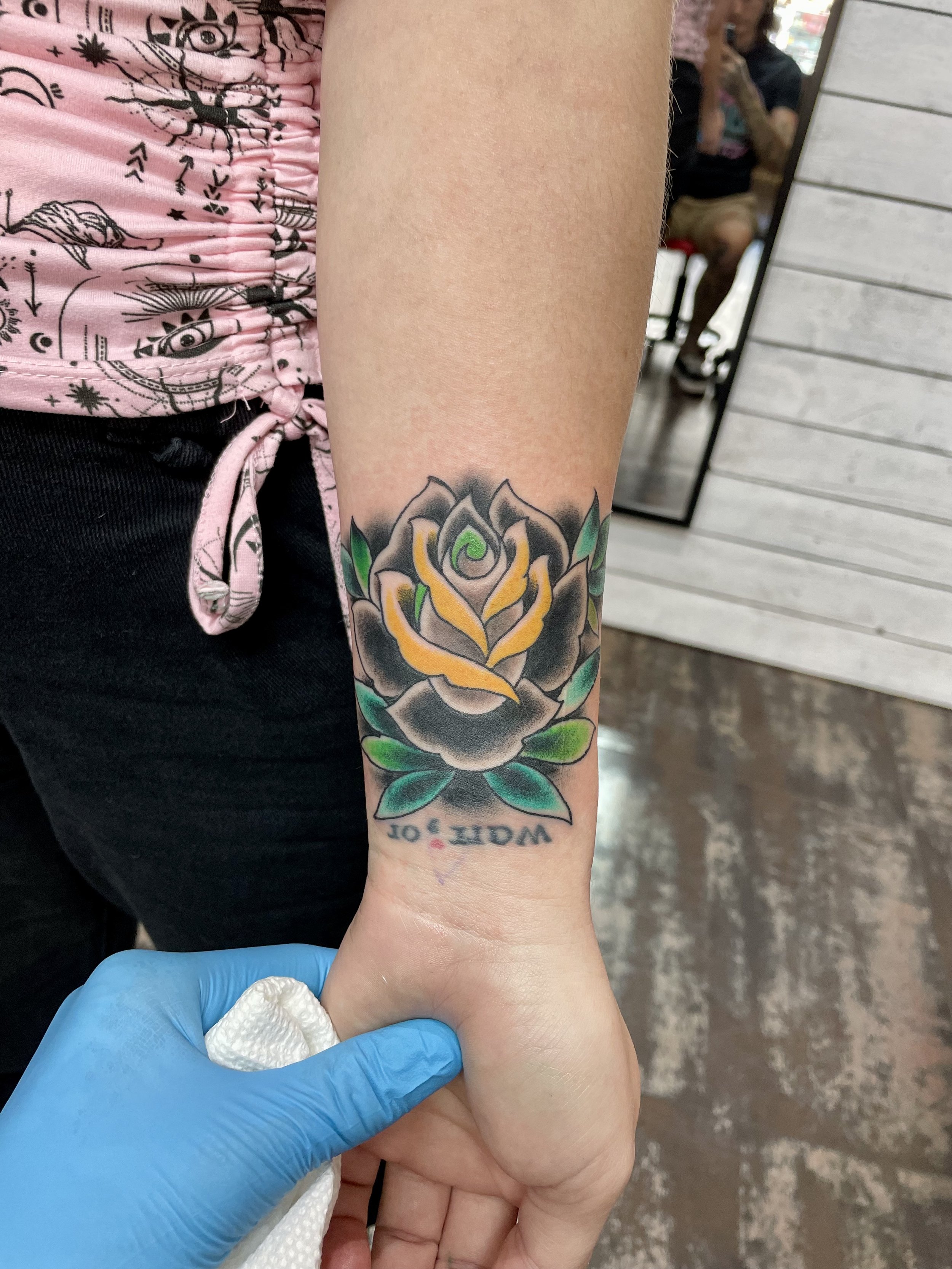 Traditional American color wrist tattoo featuring a large dark rose with bold black outer petals and a glowing gold center, surrounded by green leaves with script lettering along the lower wrist, custom tattoo by Nick at Big Kahuna Tattoo in Boca Rat