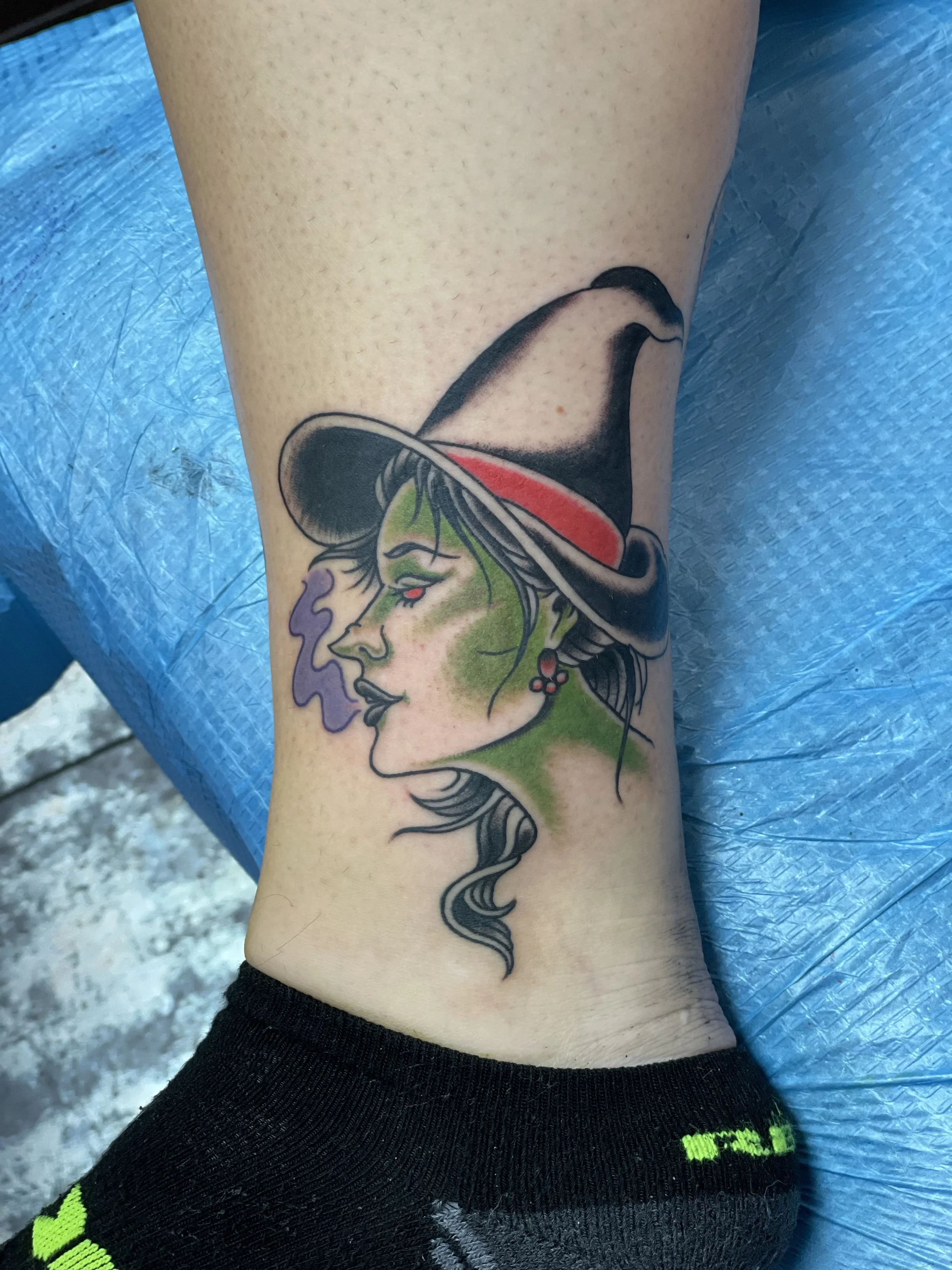 Traditional American color ankle tattoo featuring a green-skinned witch in a side profile wearing a dramatic black wide-brimmed hat with a red band, dark flowing hair, red drop earrings, and a wisp of purple smoke rising from her lips, custom tattoo 
