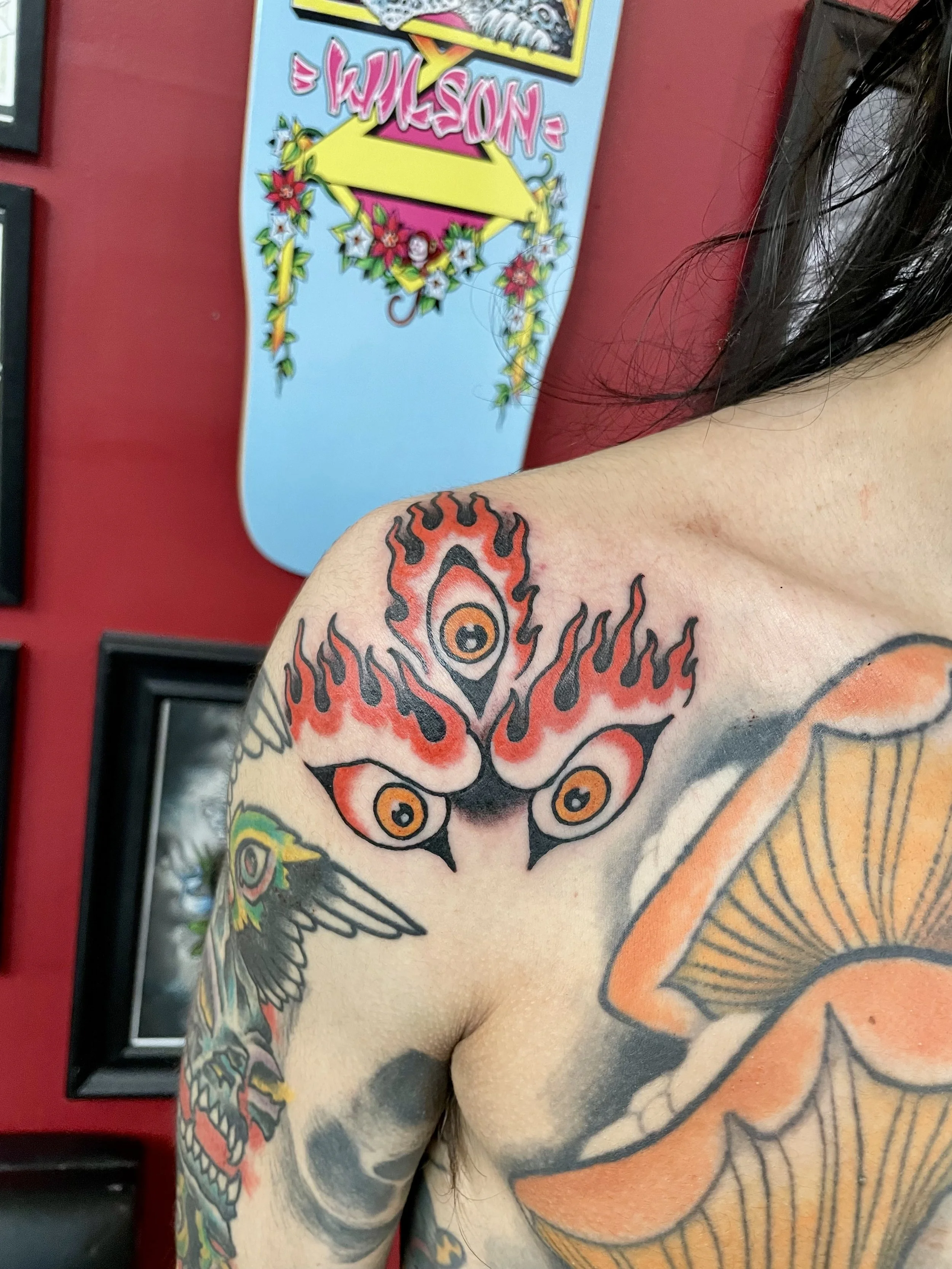Bold traditional American color shoulder tattoo featuring three orange and black flaming eyes arranged in a triangular formation, surrounded by dramatic red and black flames, set among existing sleeve work, custom tattoo by Nick at Big Kahuna Tattoo 