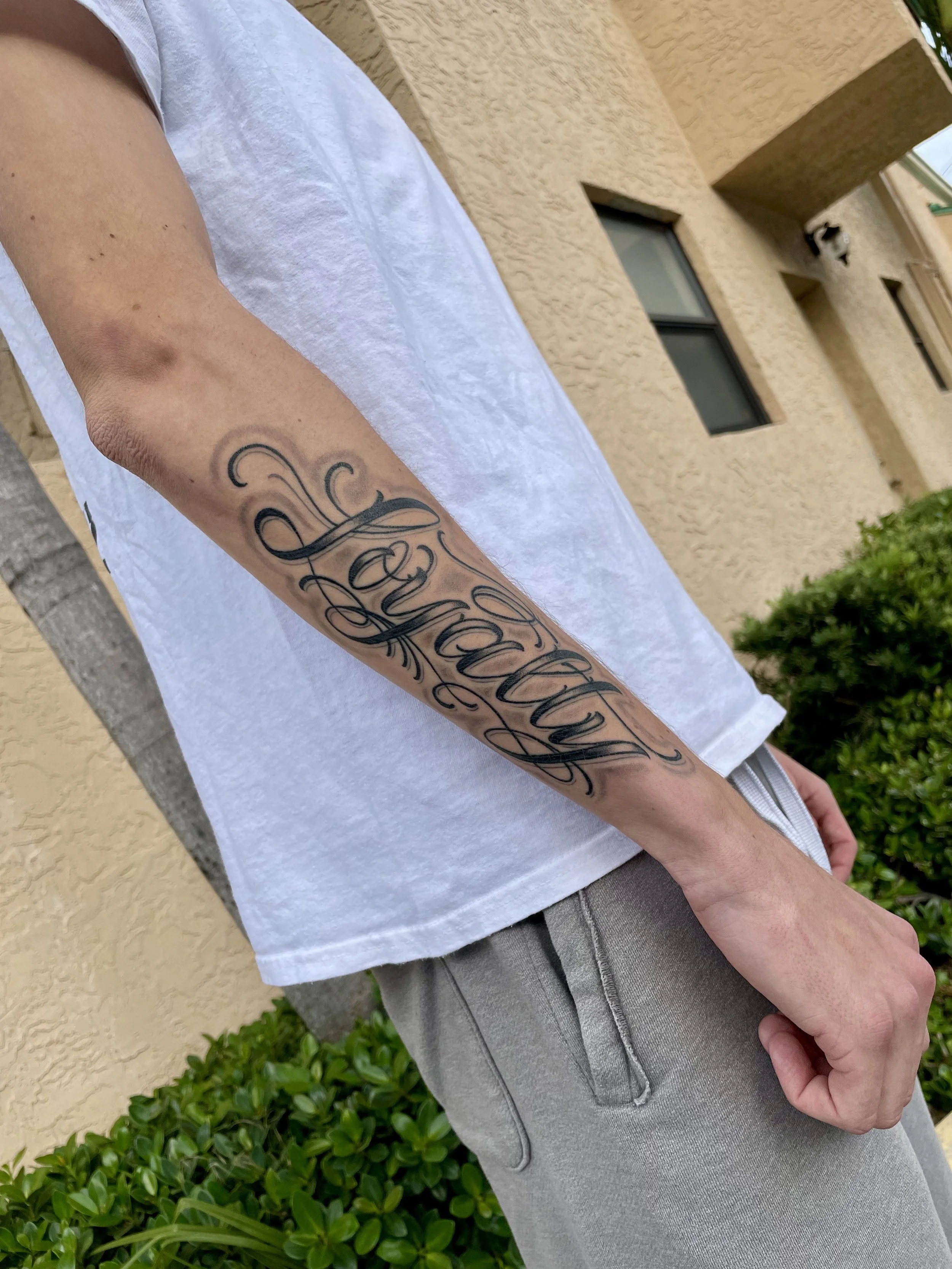 One word, said with conviction. "Loyalty" in large ornate script with sweeping flourishes above and below, spanning the full length of the forearm in bold black lettering. Black and grey forearm tattoo by Nick at Big Kahuna Tattoo in Boca Raton, FL.