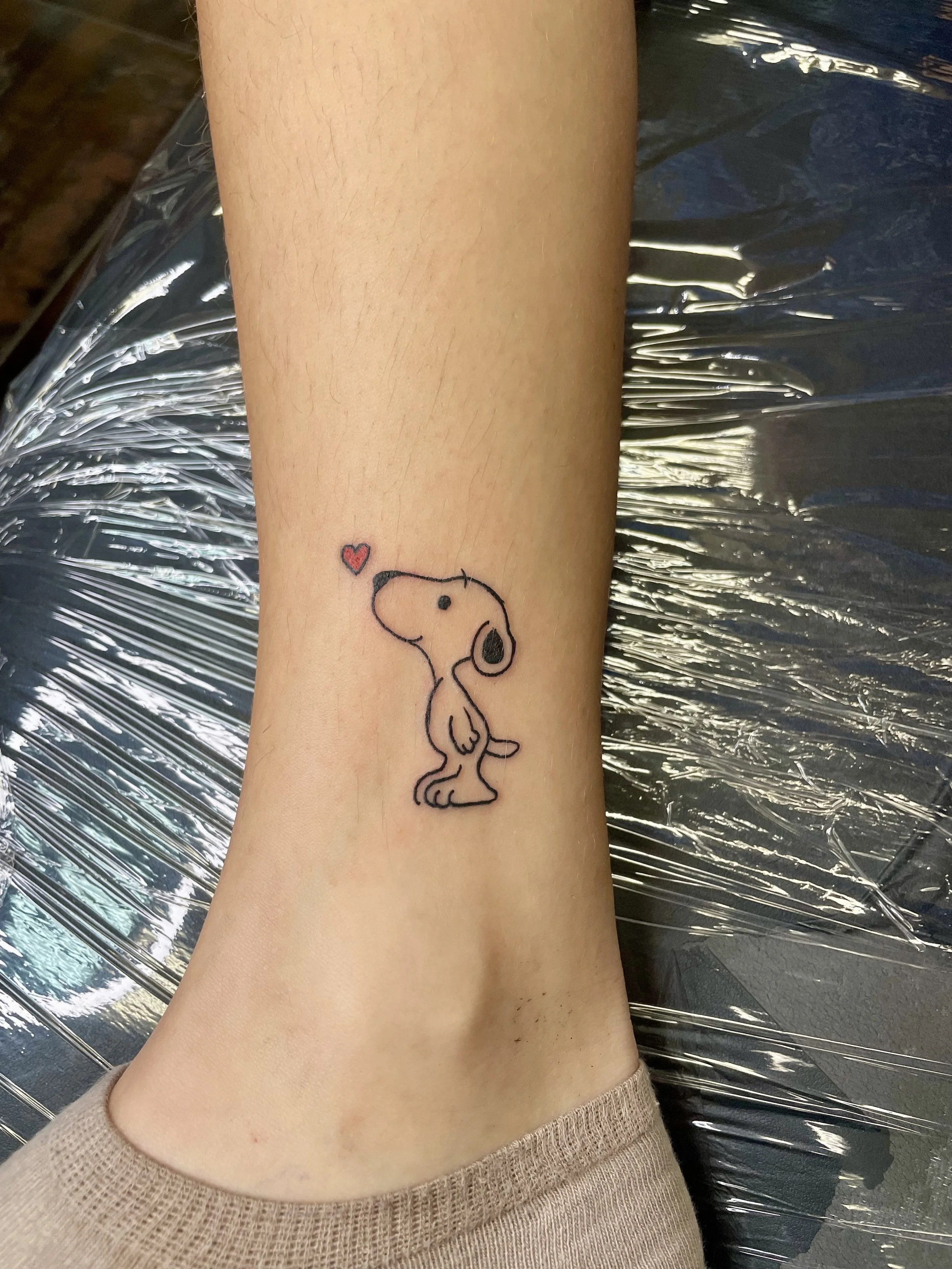 Fine line ankle tattoo featuring a sweet minimalist Snoopy in profile gazing upward at a small red heart, custom tattoo by Nick at Big Kahuna Tattoo in Boca Raton, FL.