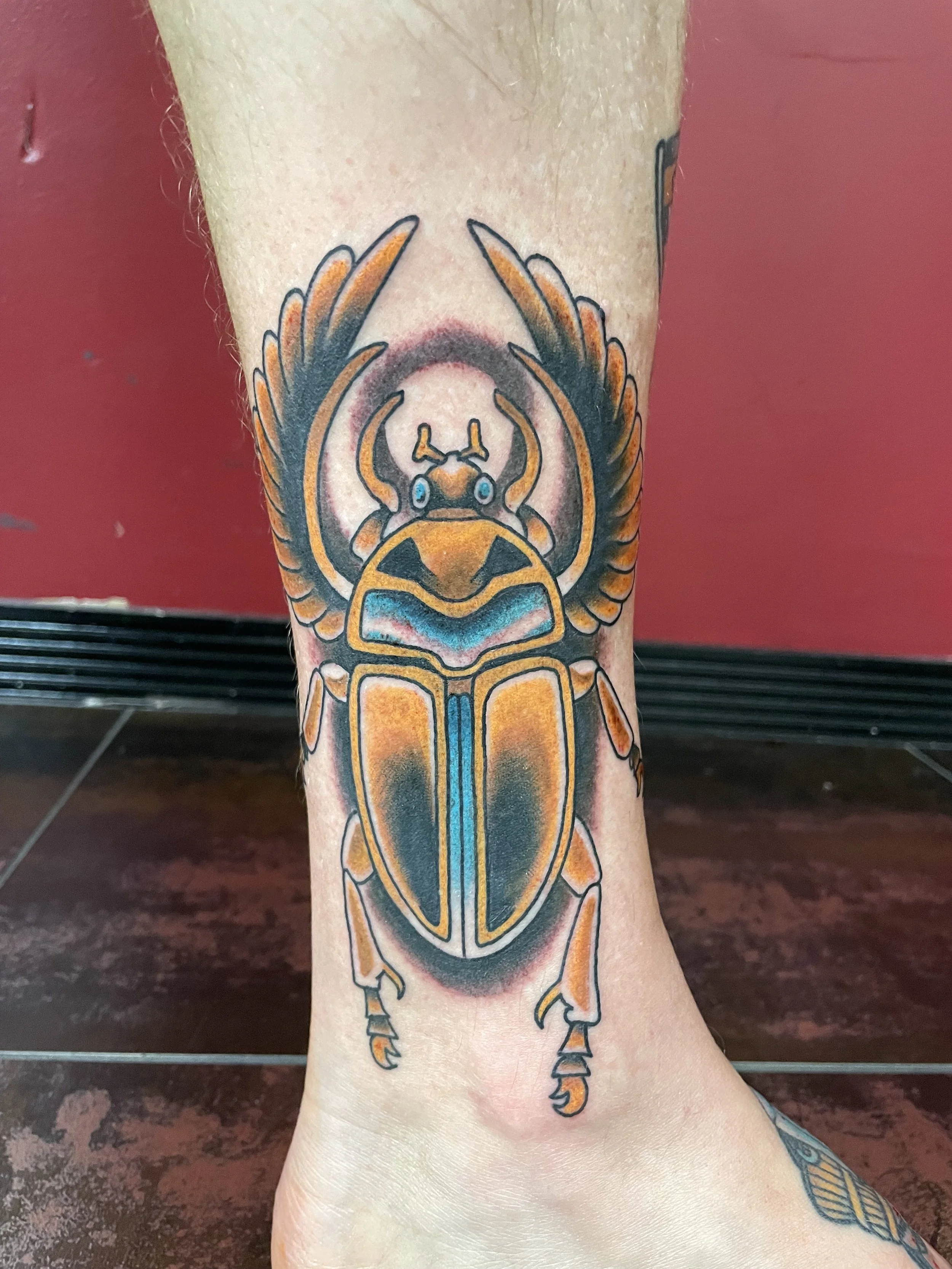 Traditional American color ankle tattoo featuring a large Egyptian scarab beetle with spread wings, rendered in rich gold and blue tones with blue eyes and detailed leg segments, custom tattoo by Nick at Big Kahuna Tattoo in Boca Raton, FL.