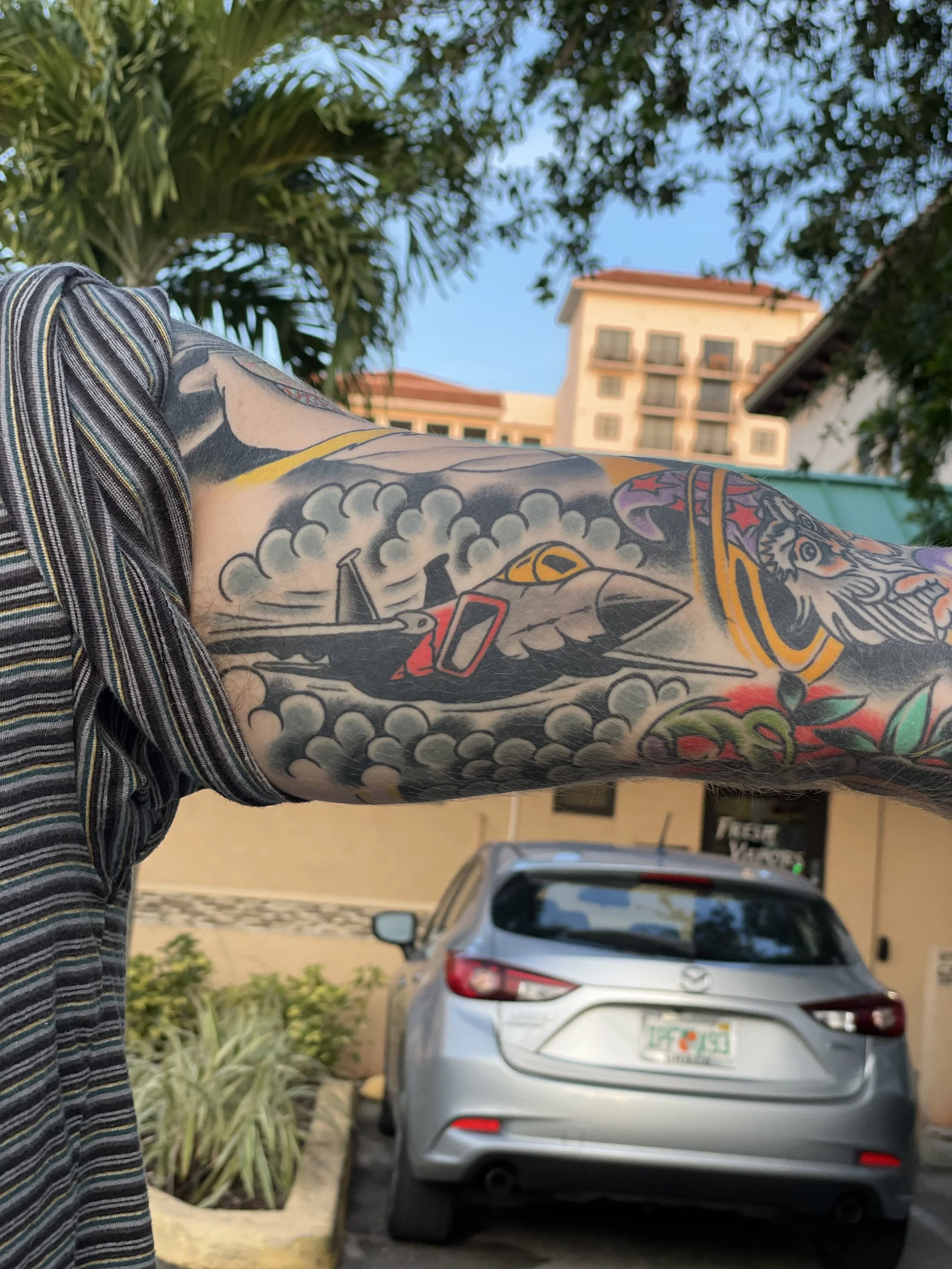 Black and grey healed forearm sleeve tattoo featuring a military fighter jet breaking through dramatic billowing clouds, set among existing color sleeve work, custom tattoo by Nick at Big Kahuna Tattoo in Boca Raton, FL.