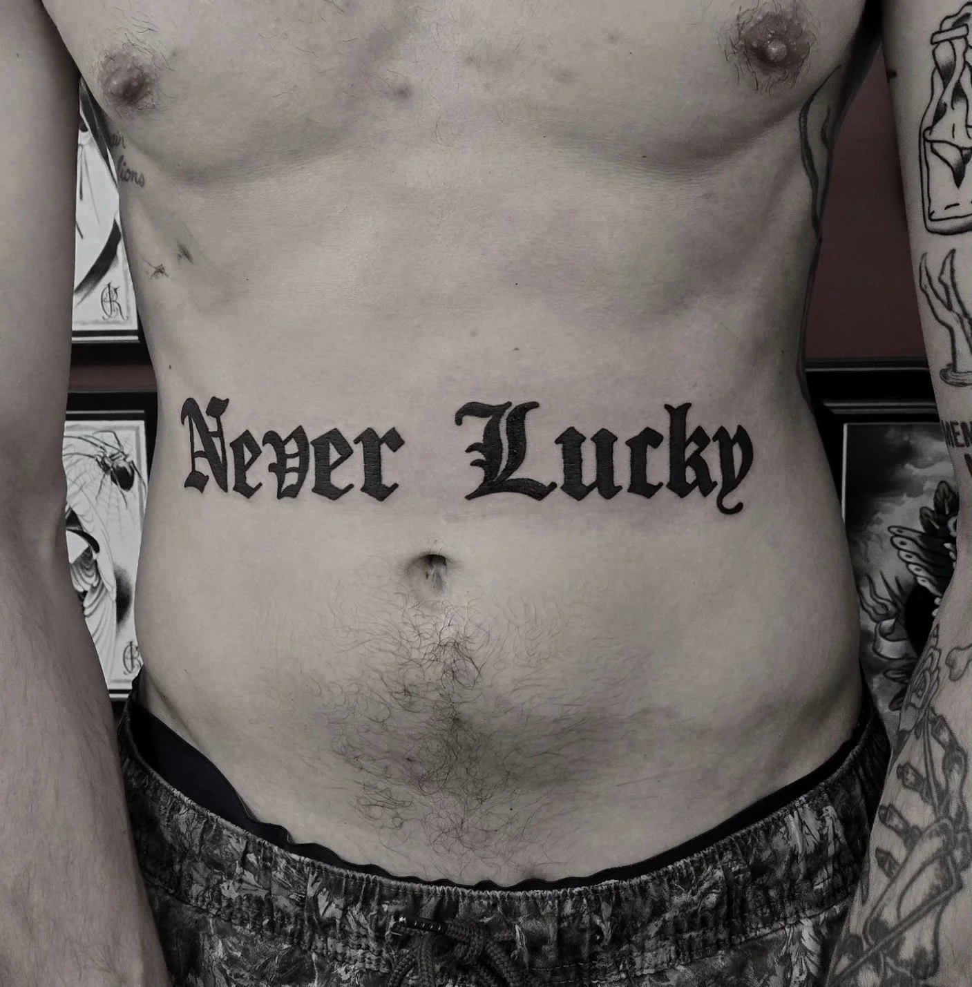 Bold black old English script stomach tattoo reading Never Lucky in large decorative lettering, custom lettering tattoo by Brandon Perez at Big Kahuna Tattoo in Boca Raton, FL