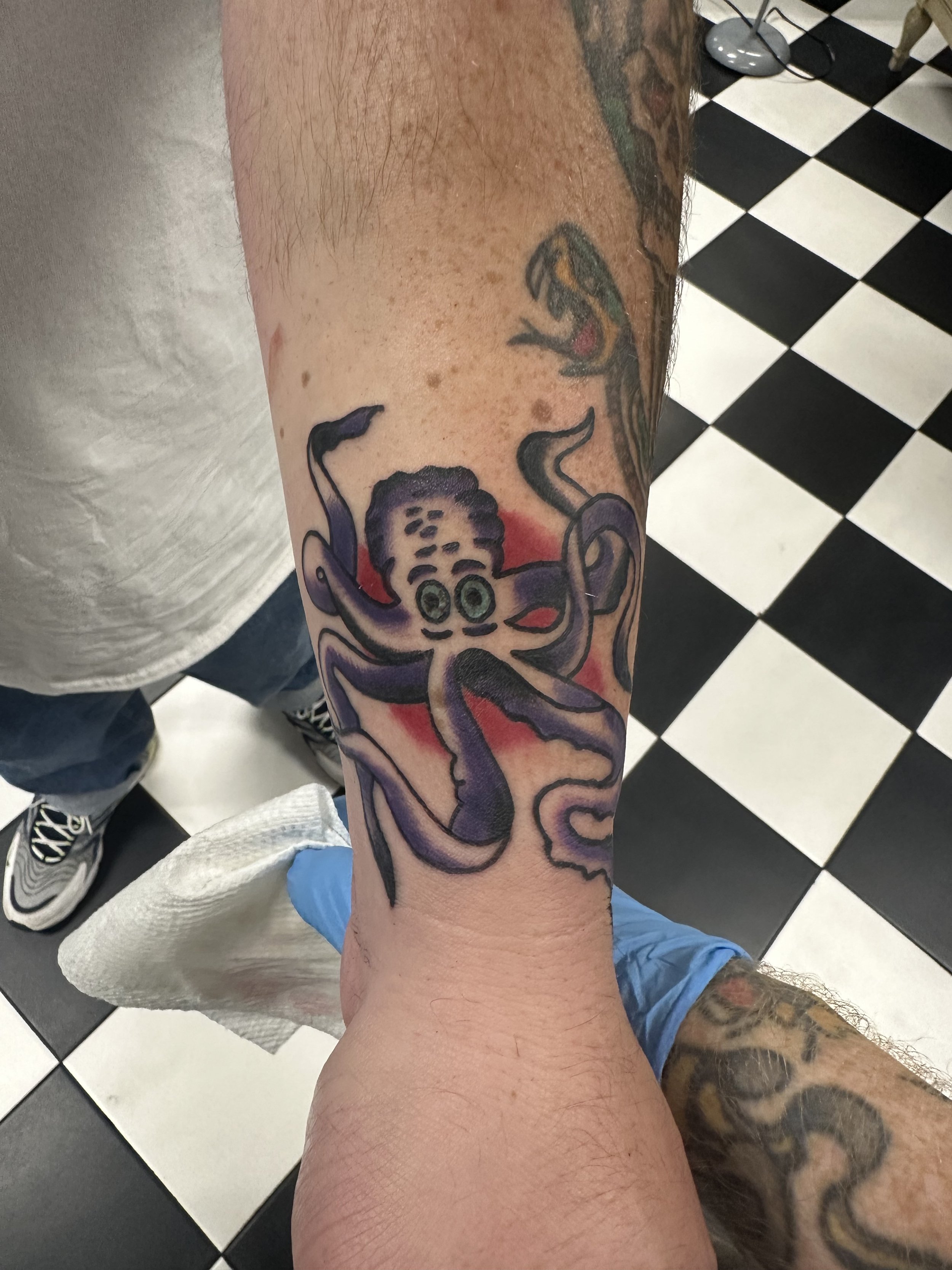 Traditional American color wrist tattoo featuring a cartoonish purple octopus with green eyes and a red background, custom tattoo by Dan at Big Kahuna Tattoo in Boca Raton, FL.