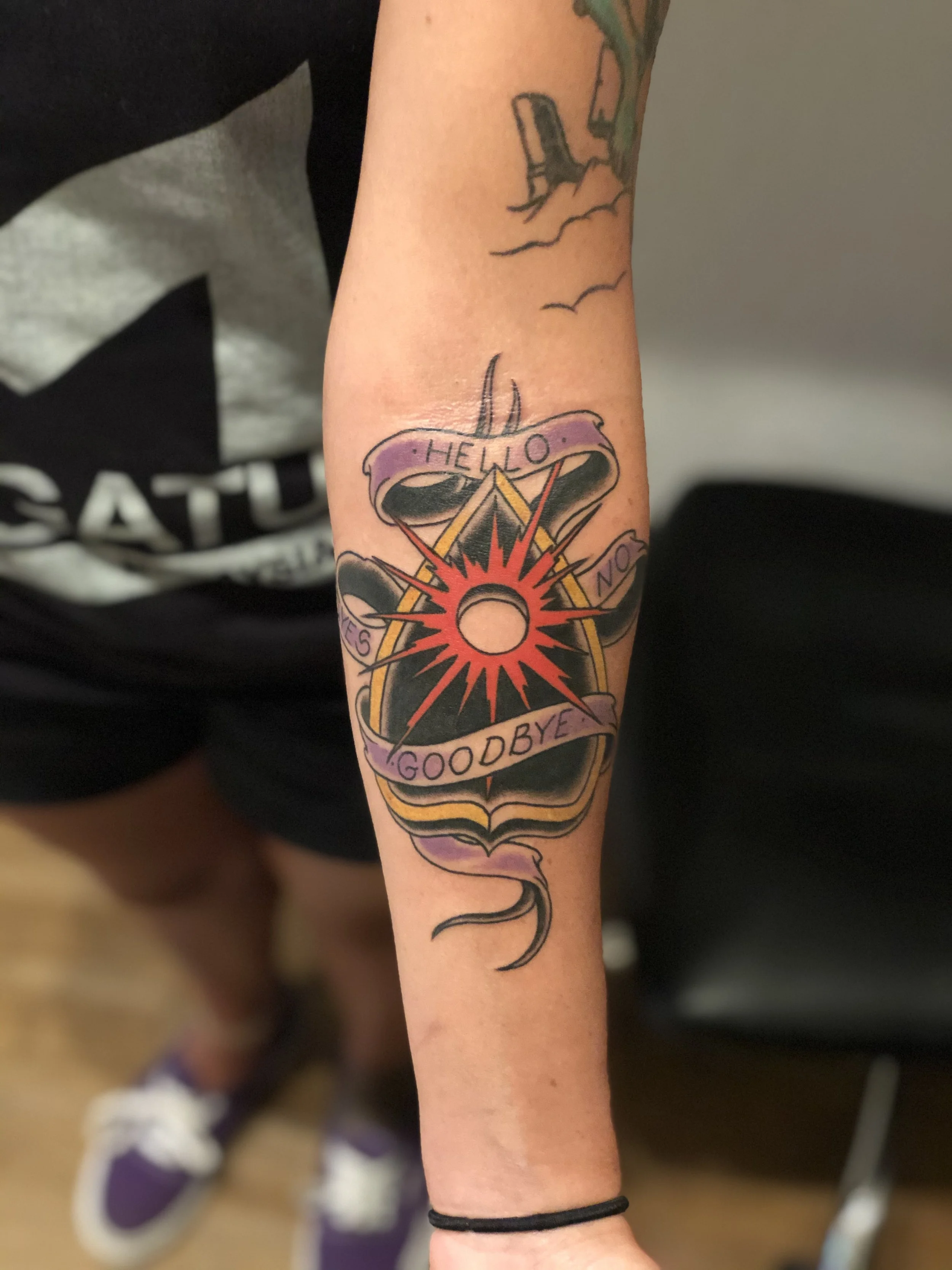 Traditional American color forearm tattoo featuring an Ouija board planchette with a red starburst and purple banners reading "Hello," "Goodbye," "Yes," and "No," custom tattoo by Nick at Big Kahuna Tattoo in Boca Raton, FL.