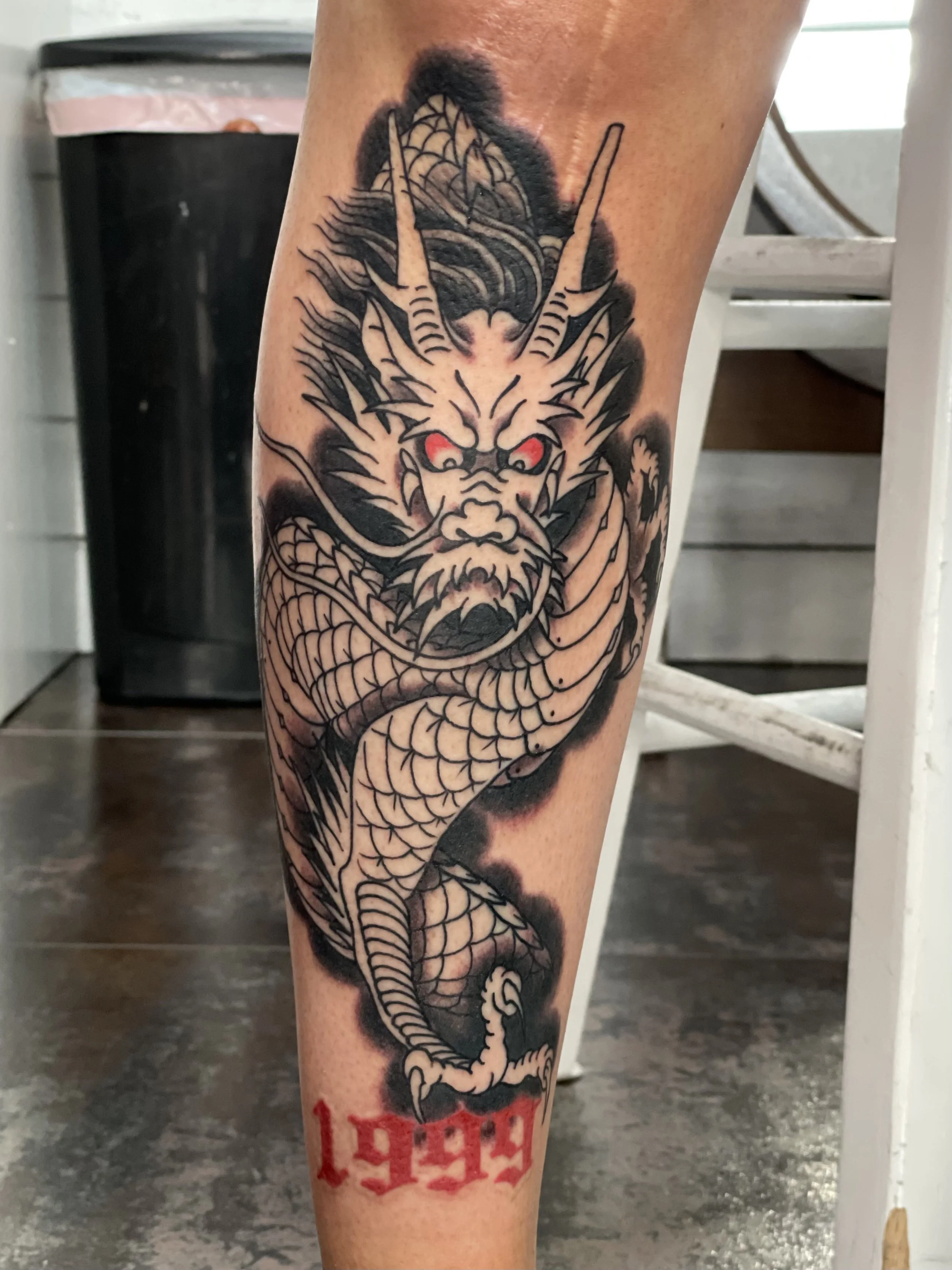 Black and grey with just two hits of red and that's all it needs. A fierce front facing Japanese dragon with glowing red eyes and a tightly coiled scaled body emerges from dark clouds, anchored by bold red "1999" lettering at the base. Shin tattoo by