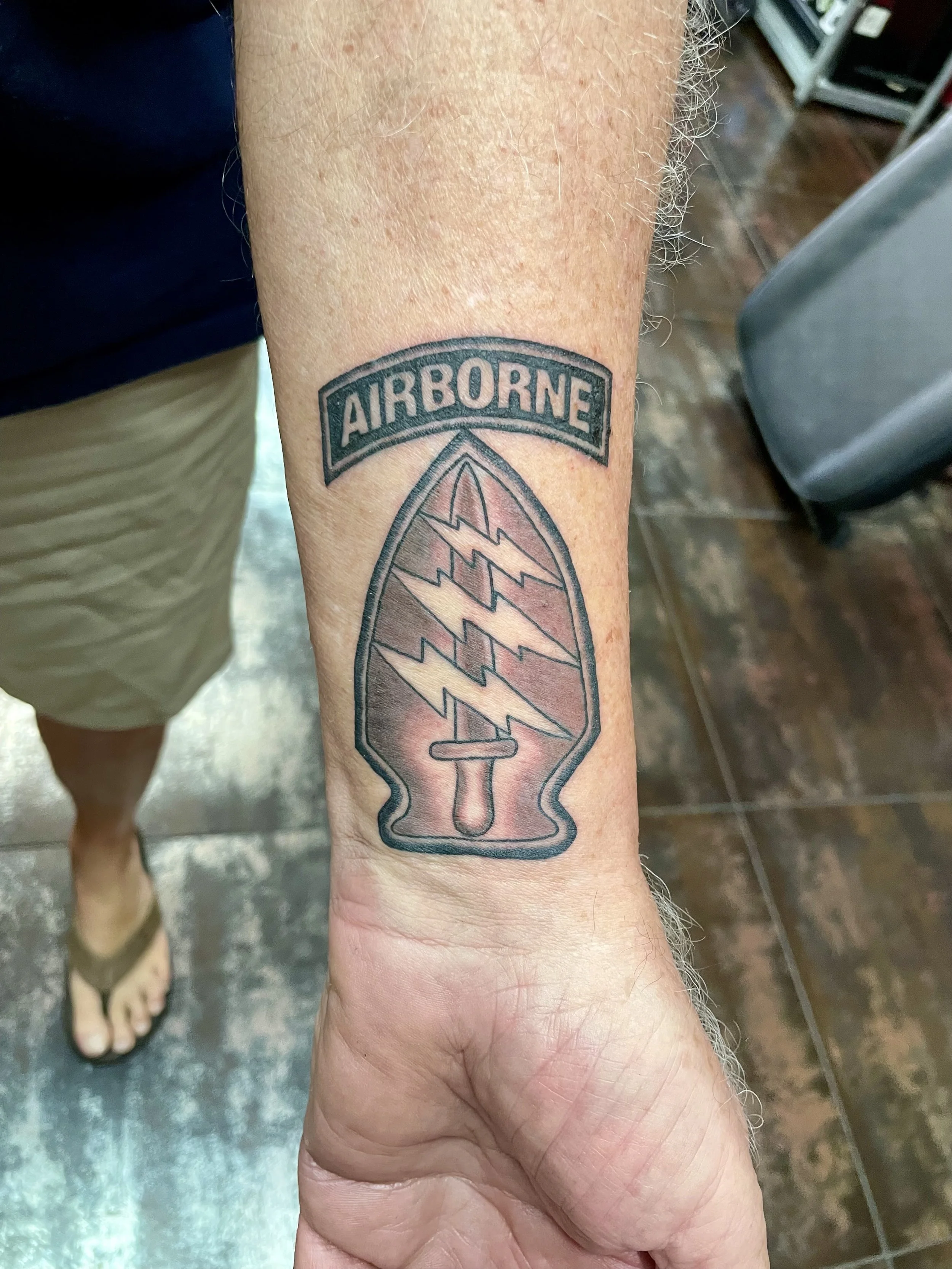 Black and grey wrist tattoo featuring the U.S. Army Special Forces arrowhead patch with crossed lightning bolts and a dagger, topped by an Airborne tab, custom tattoo by Nick at Big Kahuna Tattoo in Boca Raton, FL.
