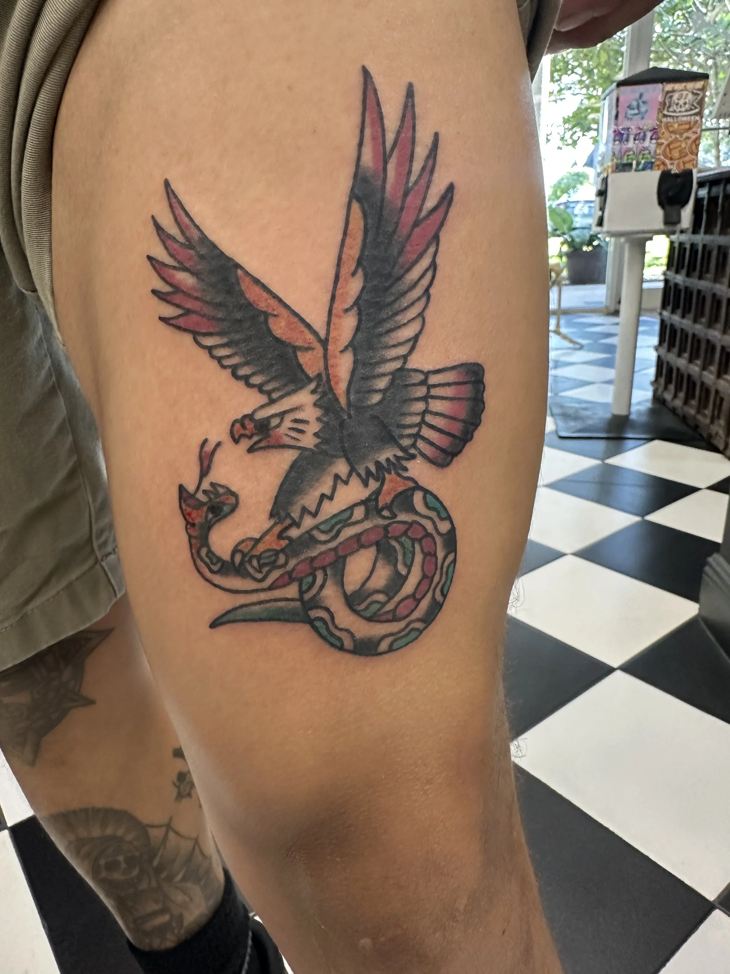 Traditional American color thigh tattoo featuring a bold bald eagle with spread red and black wings locked in battle with a coiled snake with teal and red markings, custom tattoo by Dan at Big Kahuna Tattoo in Boca Raton, FL.
