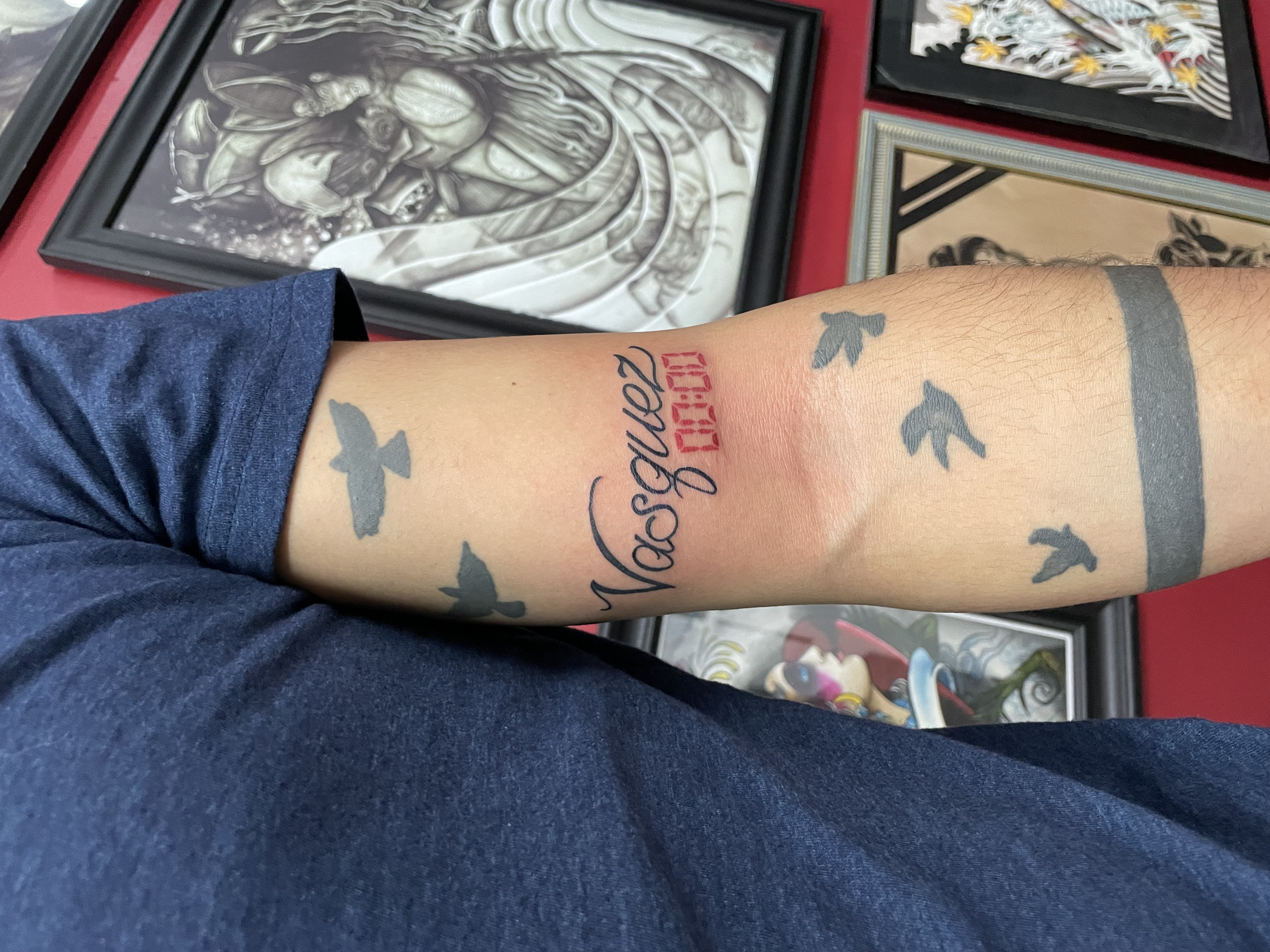 Fine line forearm tattoo featuring the name "Vasquez" in bold cursive script with a red digital clock reading 00:00 beneath it, surrounded by small silhouette birds in flight, custom tattoo by Nick at Big Kahuna Tattoo in Boca Raton, FL.