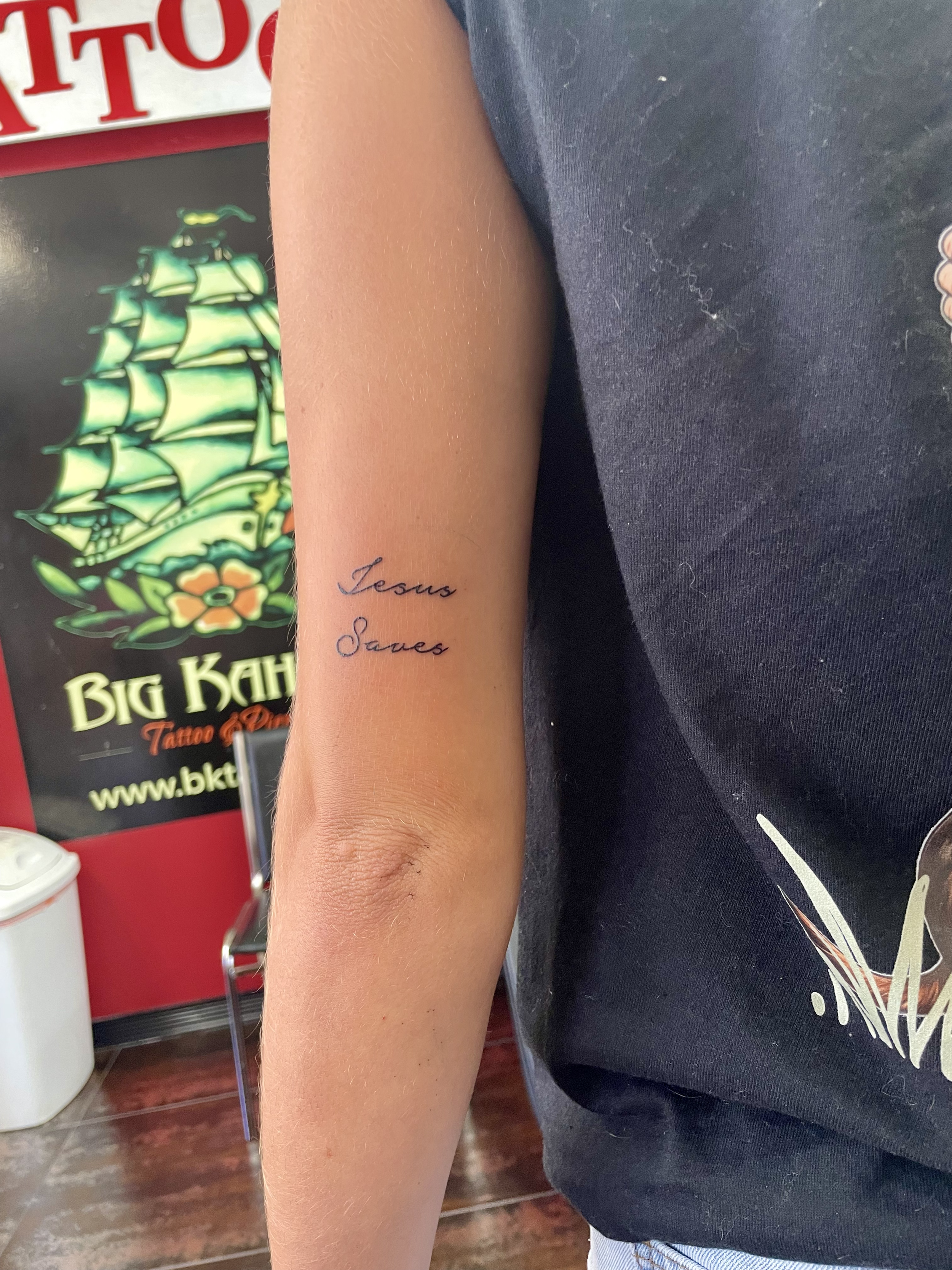 Fine line cursive script forearm tattoo reading "Jesus Saves" in elegant lettering, custom tattoo by Nick at Big Kahuna Tattoo in Boca Raton, FL.