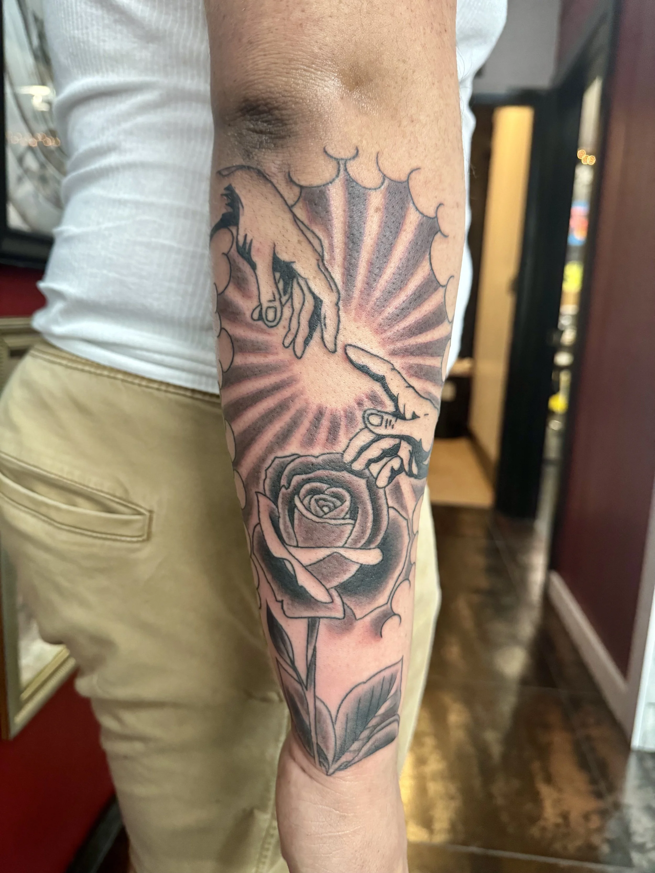 Black and grey forearm tattoo featuring the outstretched hands from Michelangelo's Creation of Adam reaching toward each other with radiating light beams between them, above a large traditional rose with stem and leaves, custom tattoo by Nick at Big 