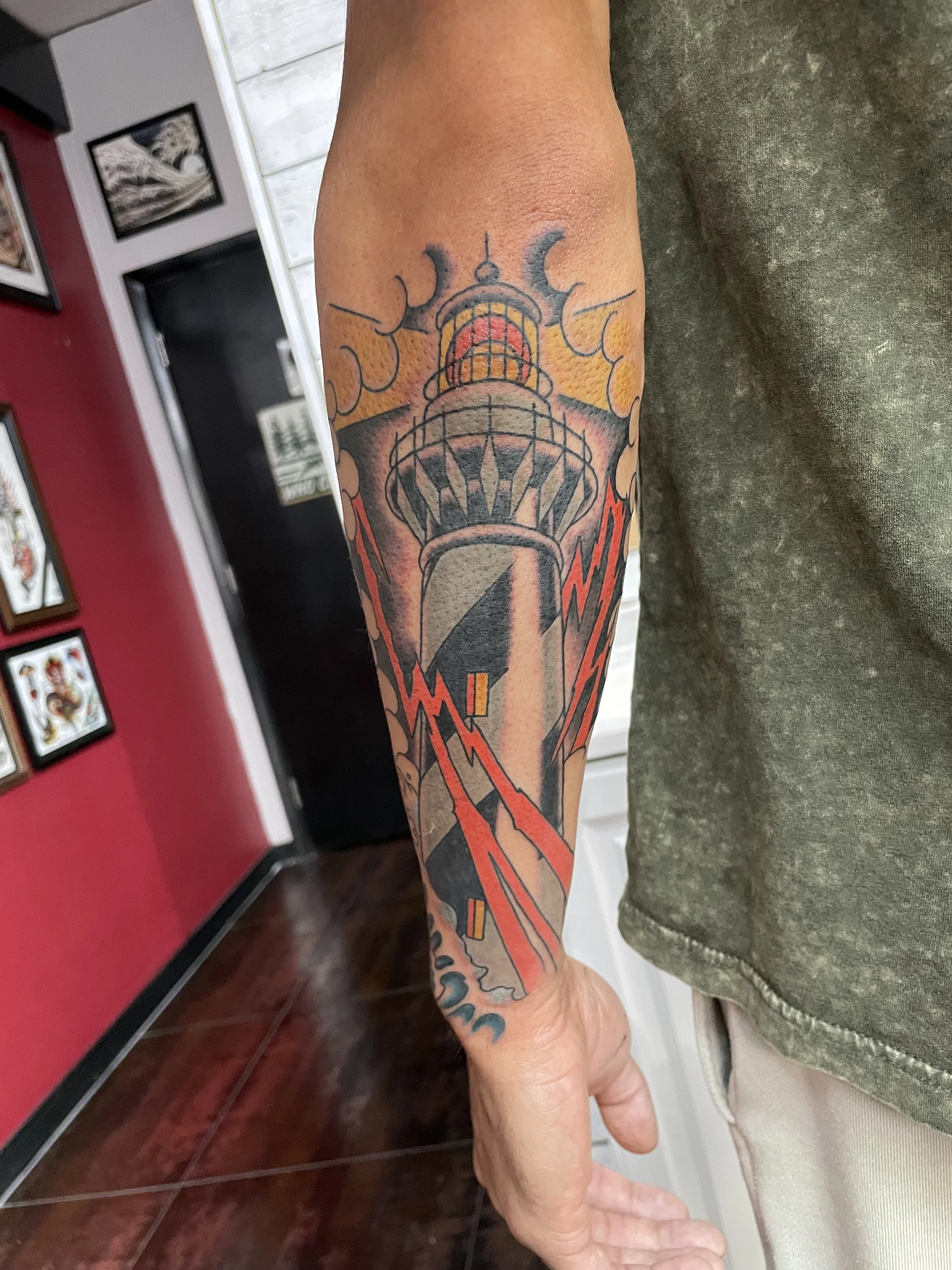 A lighthouse getting hammered by a storm with red lightning bolts from every direction, the beacon still glowing warm orange and red at the top. Color forearm tattoo by Nick at Big Kahuna Tattoo in Boca Raton, FL.