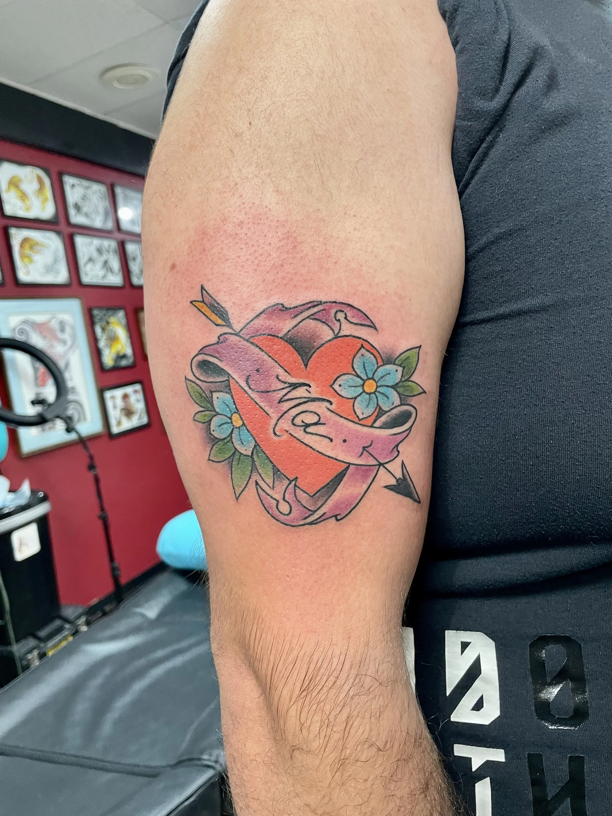 Traditional American color forearm tattoo featuring a bold red heart pierced by an arrow and wrapped in a flowing purple banner, flanked by blue forget-me-not flowers and green leaves, custom tattoo by Nick at Big Kahuna Tattoo in Boca Raton, FL.