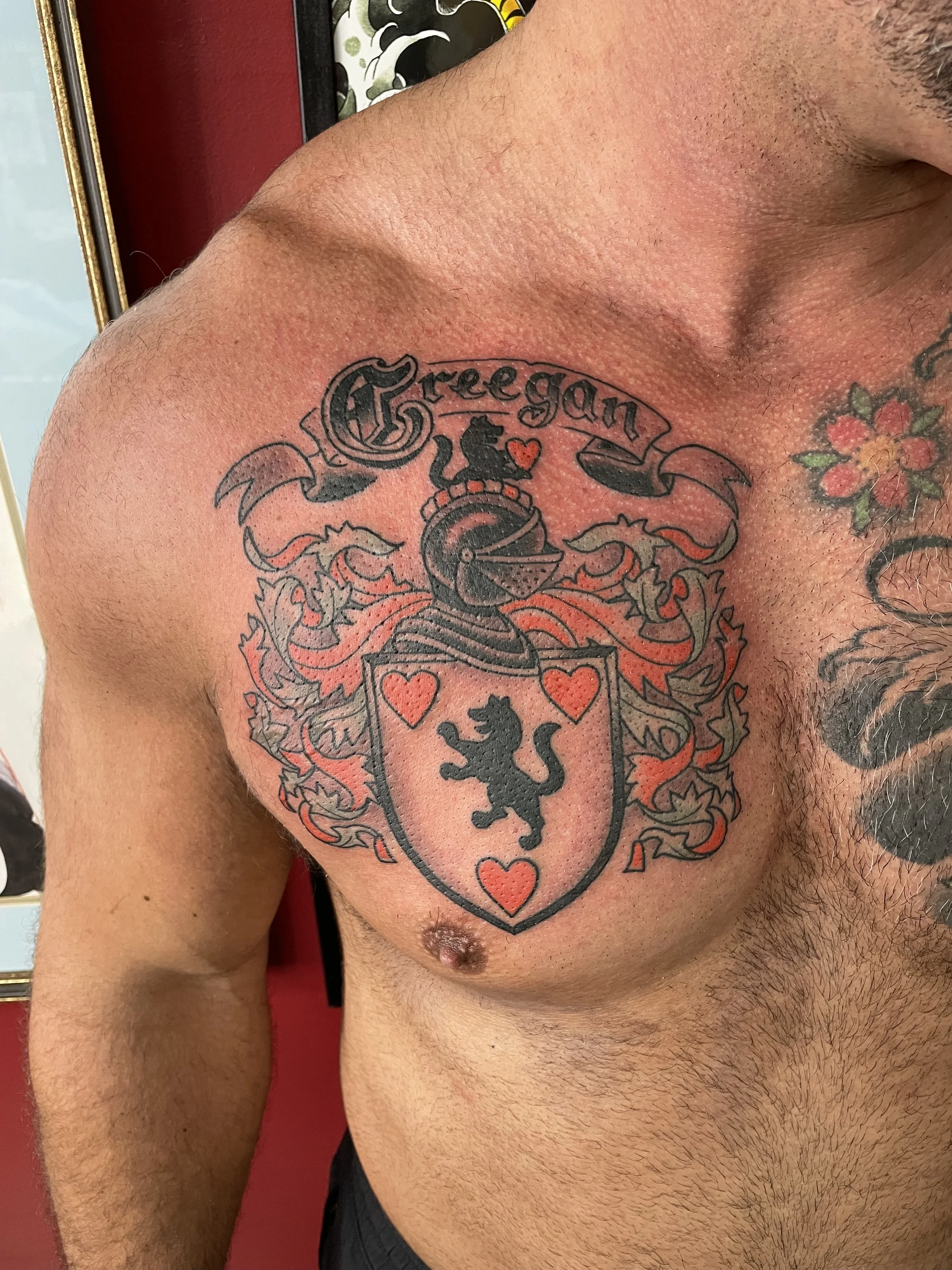 Traditional American black and grey chest tattoo featuring the Creegan family coat of arms with a shield displaying a rampant lion and three hearts, topped by a crested helmet with flowing mantling, framed by a scroll banner bearing the family name, 