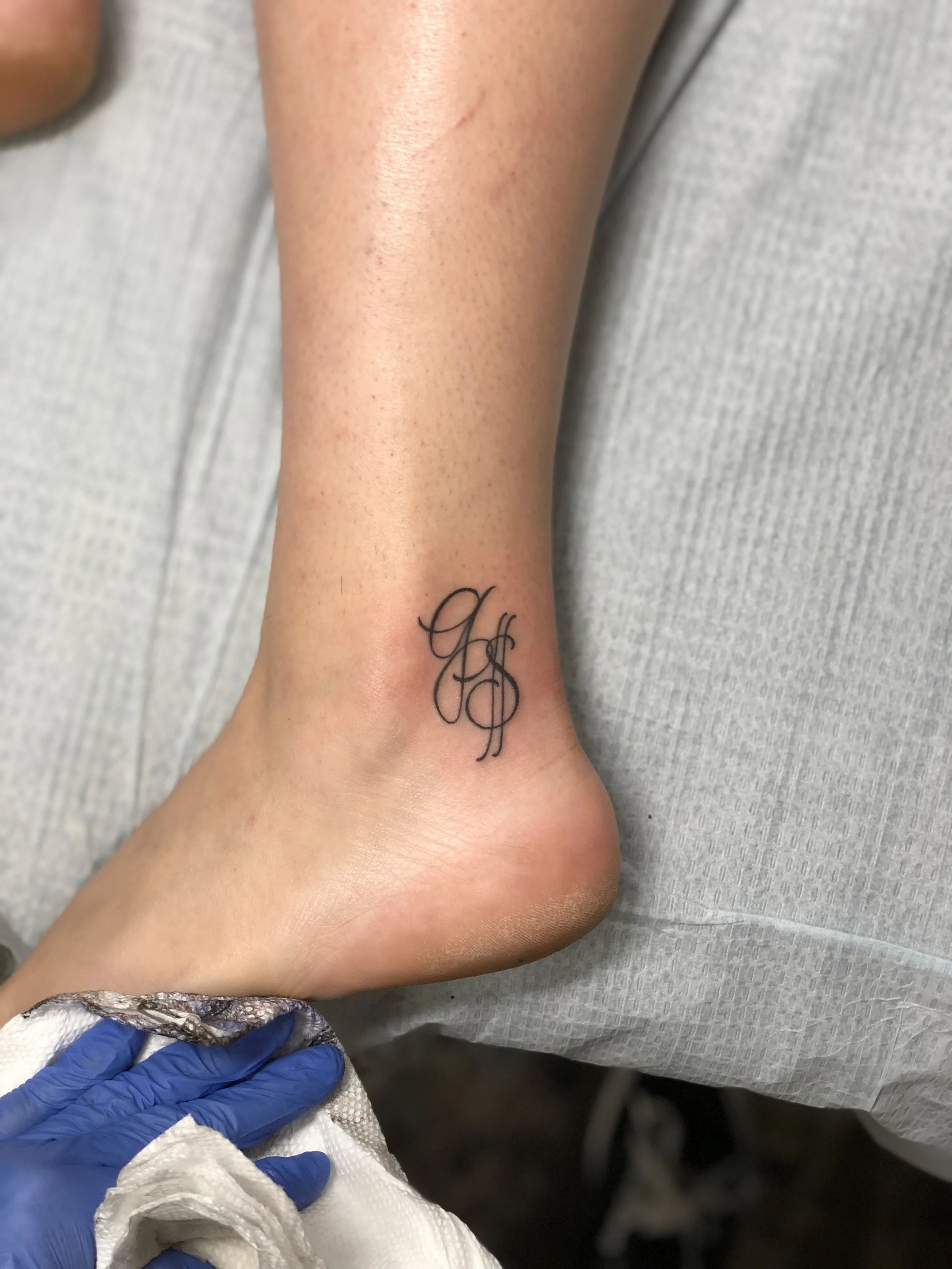 Delicate fine line ankle tattoo featuring an elegant interlocking monogram in ornate cursive script, custom tattoo by Nick at Big Kahuna Tattoo in Boca Raton, FL.