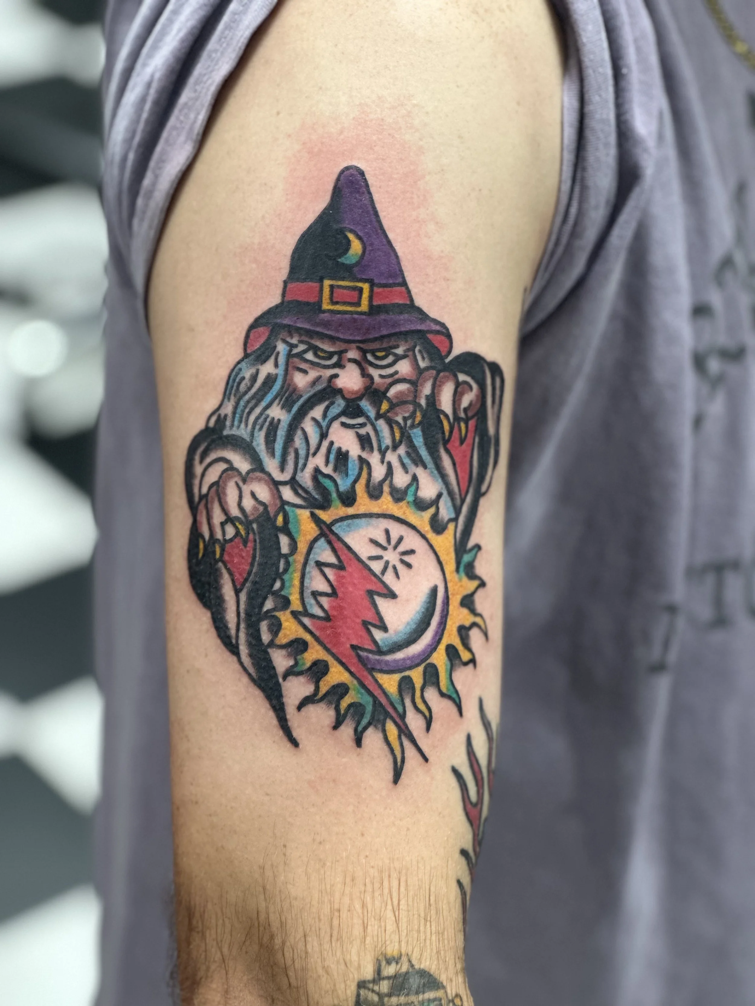 Traditional American color upper arm tattoo featuring a scowling wizard in a purple hat with a moon symbol, clutching a glowing sun and crescent moon orb with a red lightning bolt, custom tattoo by Dan at Big Kahuna Tattoo in Boca Raton, FL.