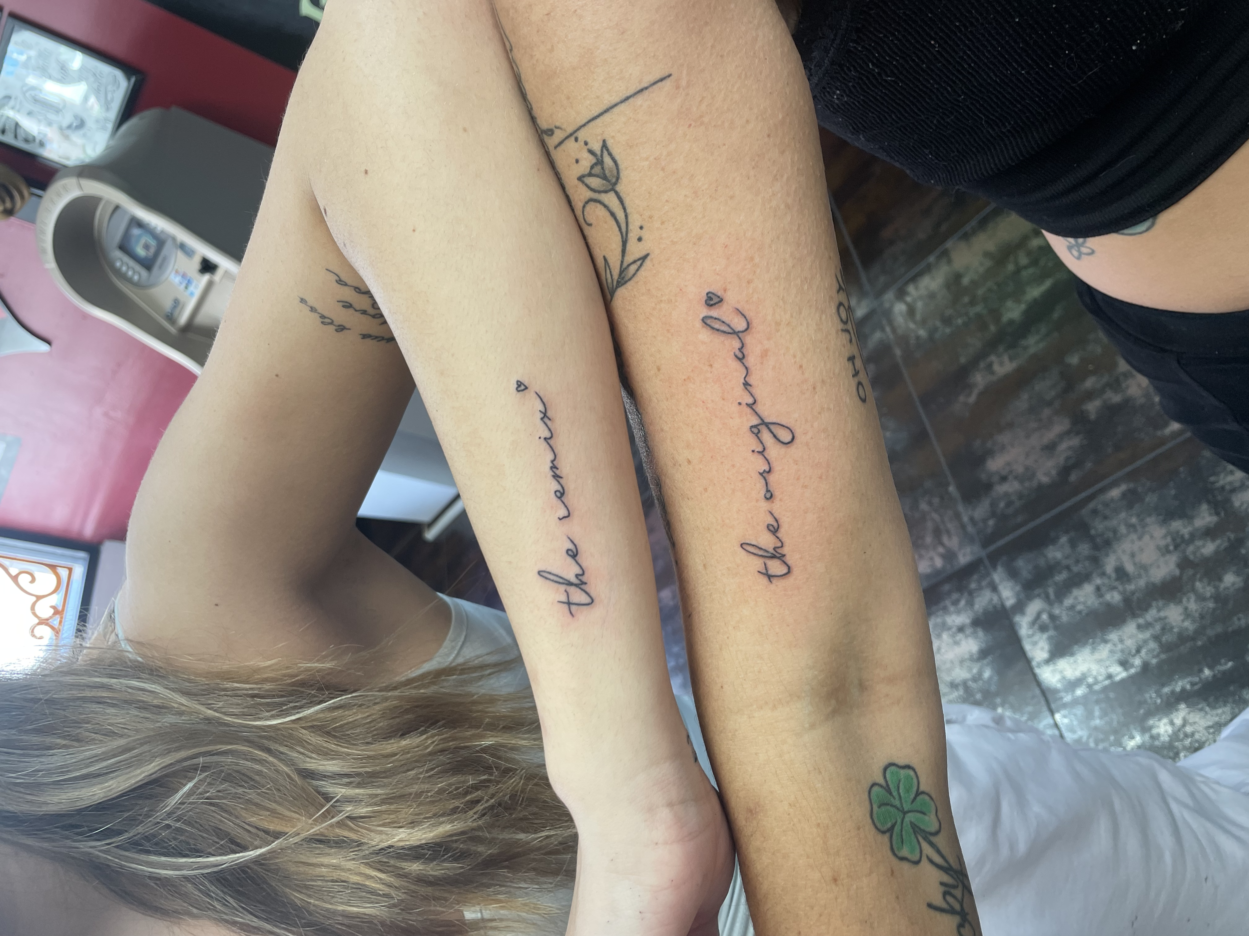Matching fine line cursive forearm tattoos for a mother and daughter set, one reading "the original" and the other "the remix" each with a small heart, custom tattoos by Nick at Big Kahuna Tattoo in Boca Raton, FL.