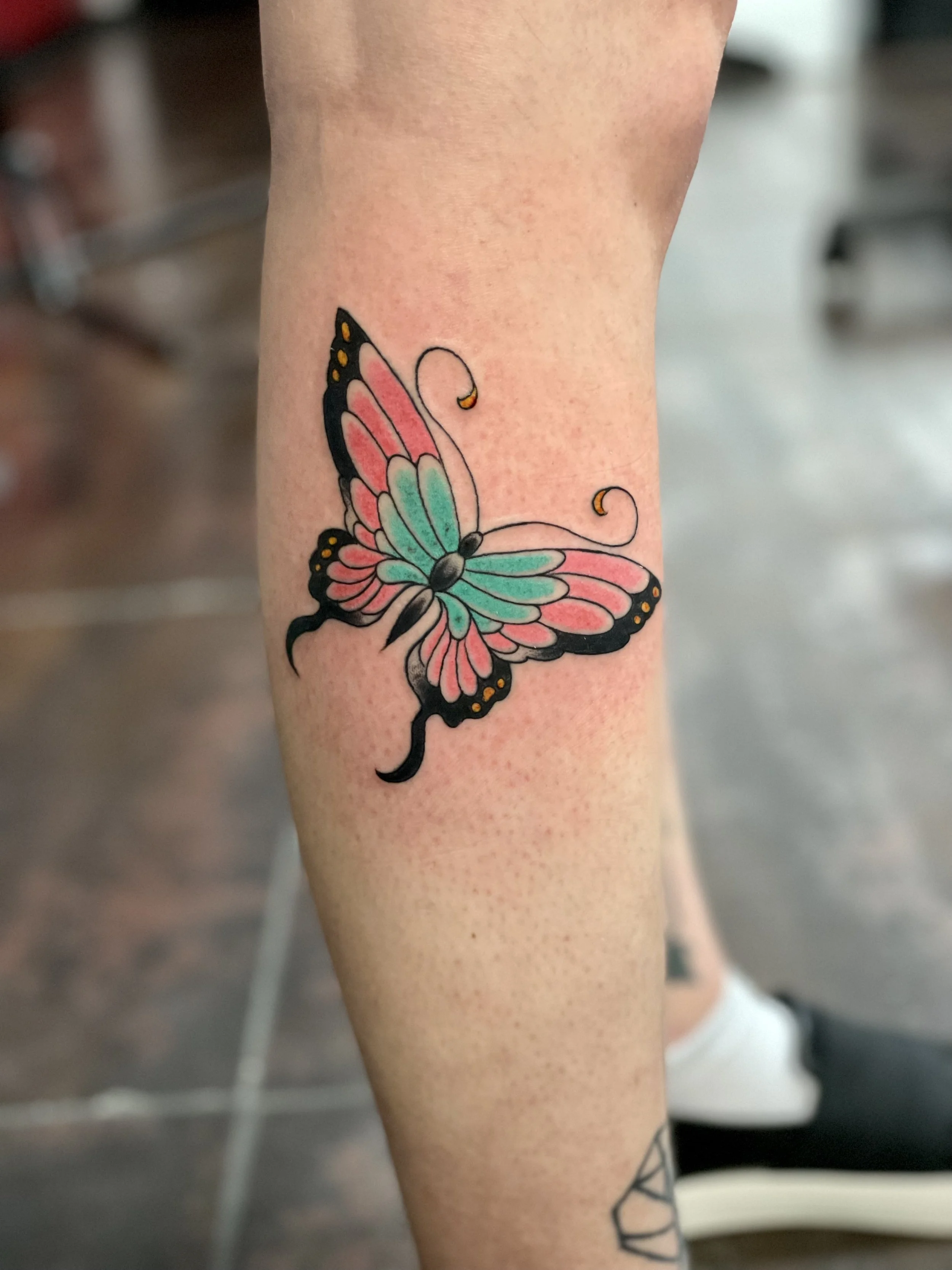 Traditional Fine Line American color calf tattoo featuring a bold butterfly with pink and teal wings, black outlined borders, and gold dot accents with decorative curling antennae, custom tattoo by Nick at Big Kahuna Tattoo in Boca Raton, FL.