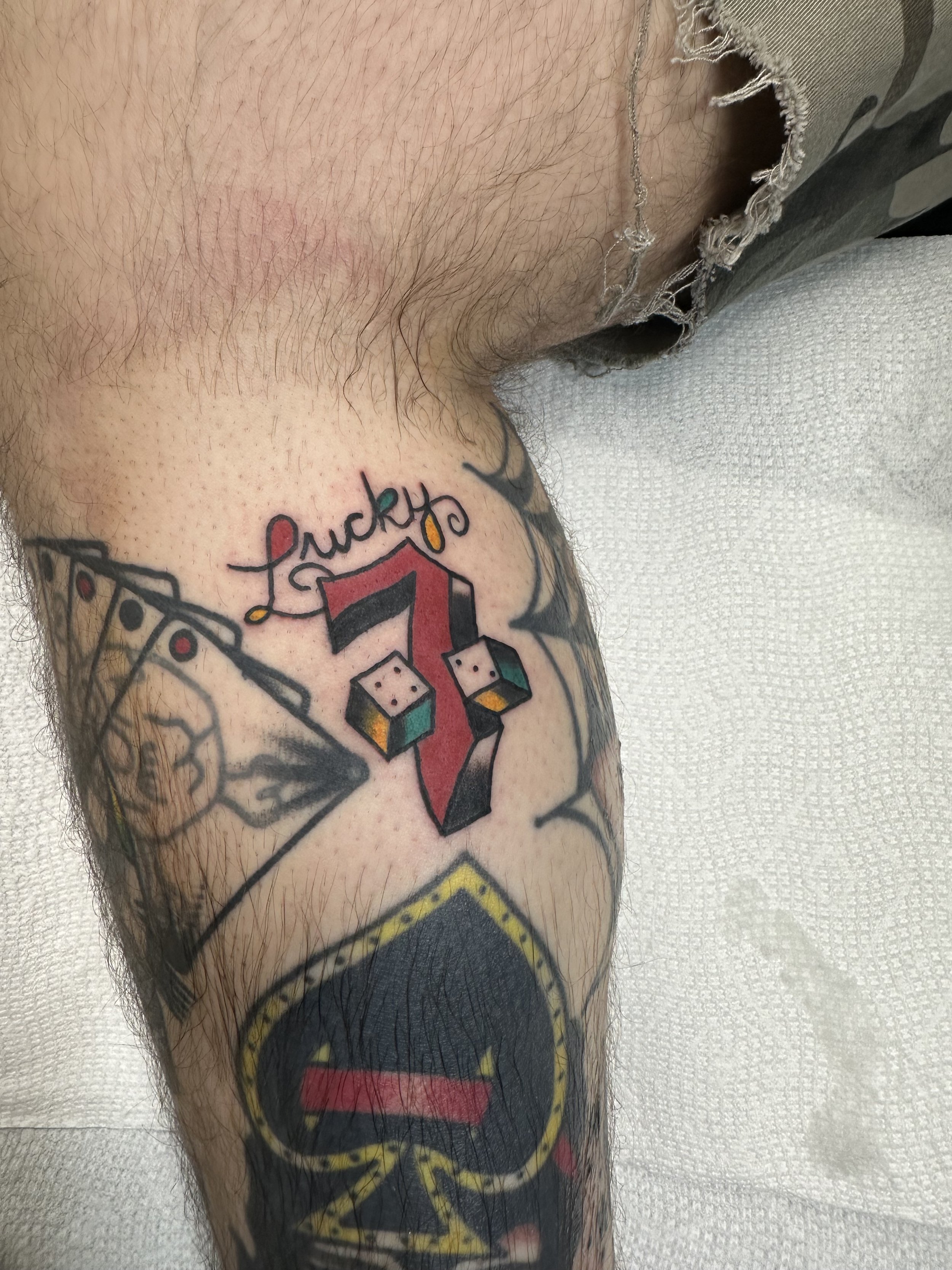 
Traditional American color leg tattoo featuring a bold red Lucky 7 with stacked dice at the base and cursive "Lucky" script above, part of a larger gambling-themed leg sleeve, custom tattoo by Dan at Big Kahuna Tattoo in Boca Raton, FL.