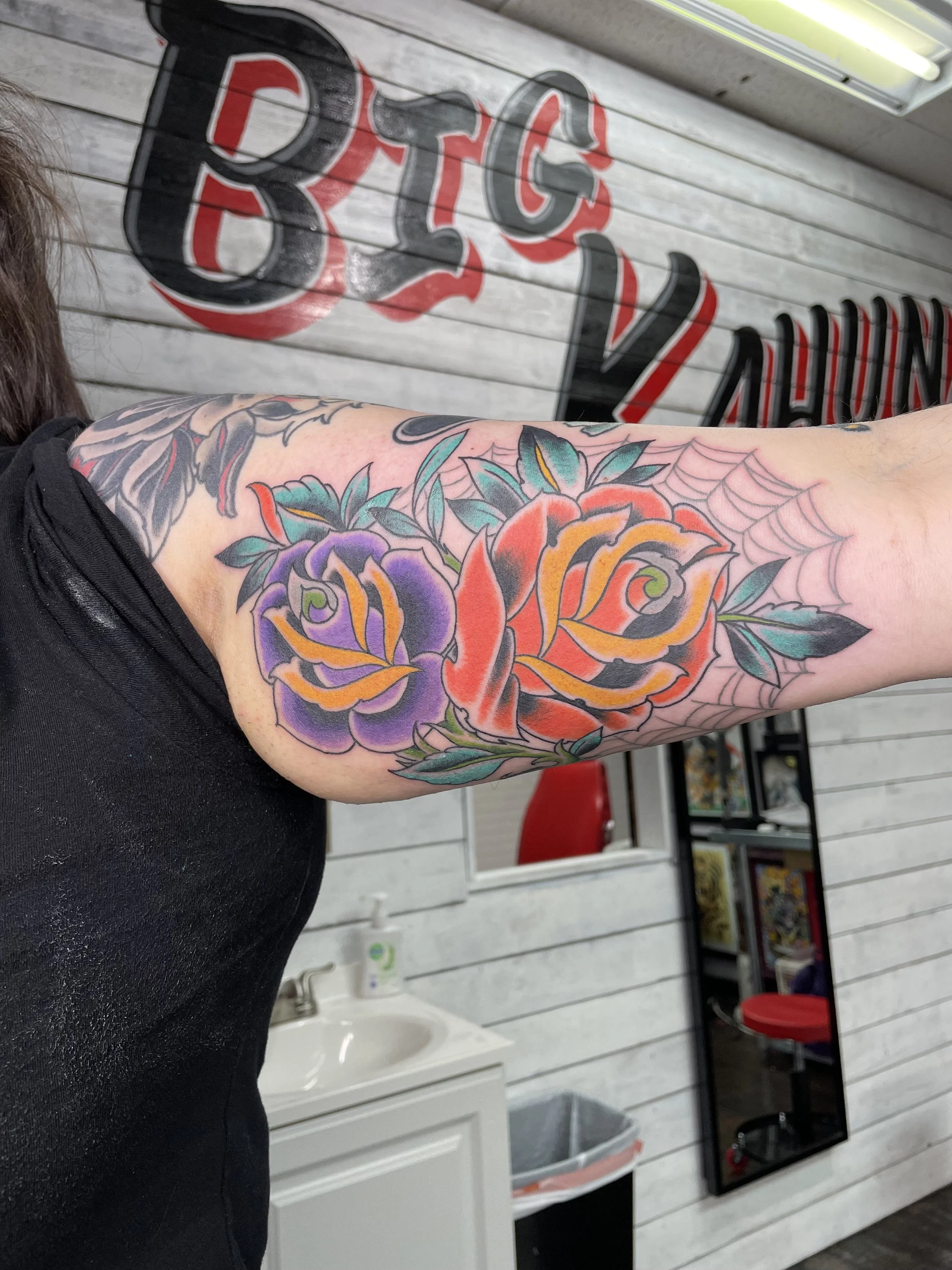 Traditional American color upper arm tattoo featuring two large blooming roses in purple and red-orange with teal foliage, set alongside a classic spiderweb elbow design, custom tattoo by Nick at Big Kahuna Tattoo in Boca Raton, FL.