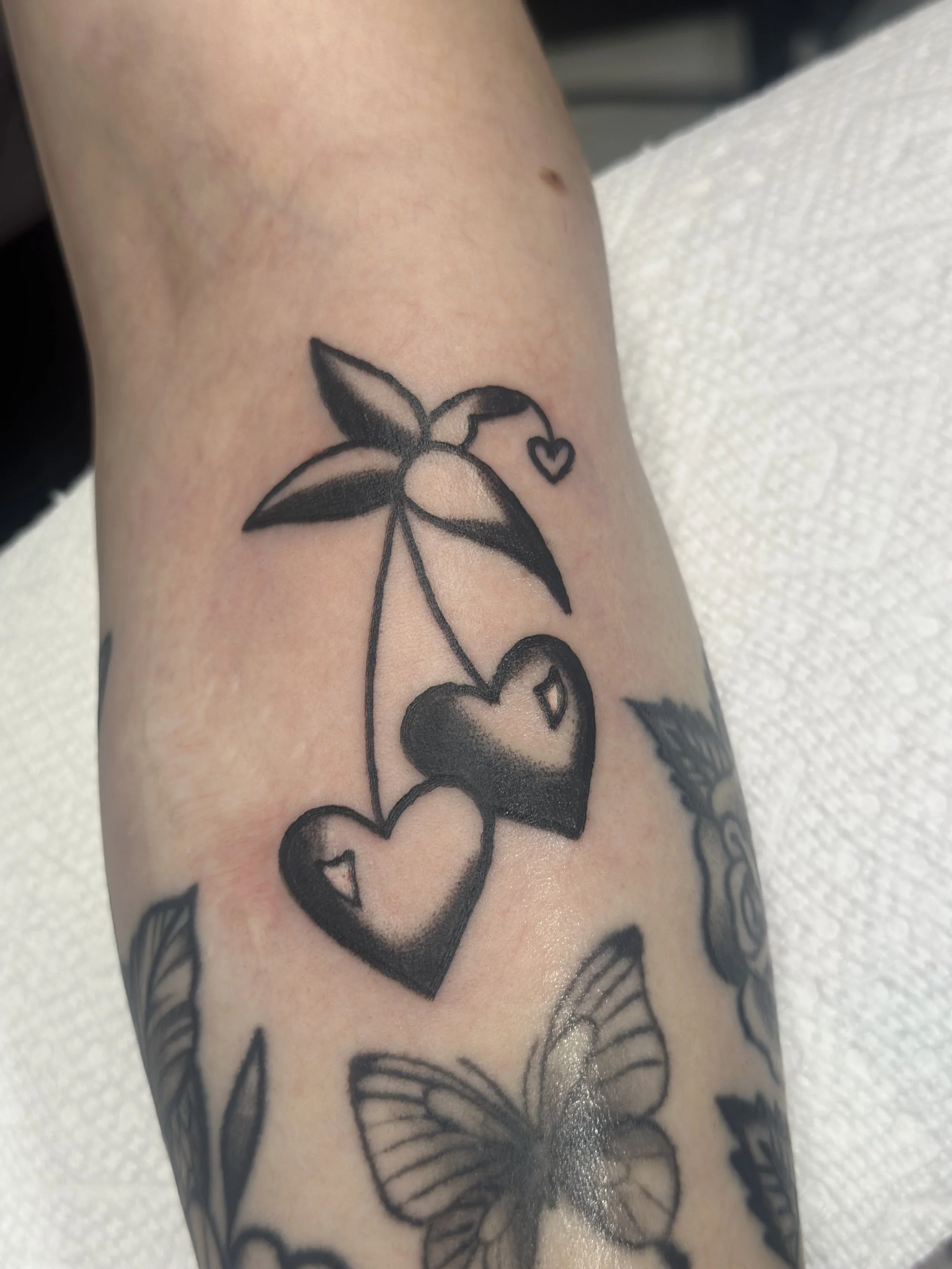 Traditional American black and grey forearm tattoo featuring a bold cherry design with heart-shaped fruit and a small floating heart, part of a larger floral sleeve, custom tattoo by Dan at Big Kahuna Tattoo in Boca Raton, FL.
