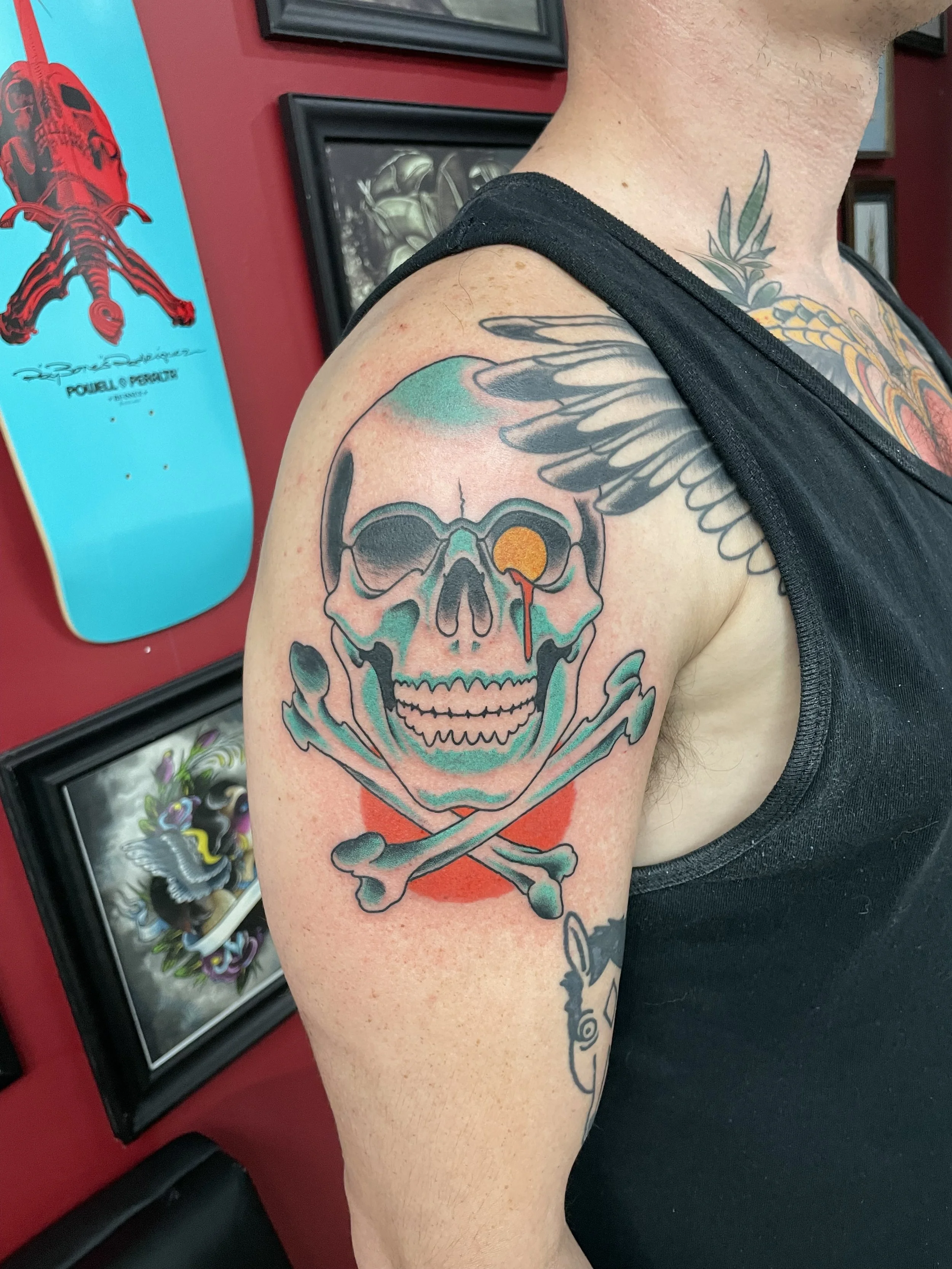 Traditional American color upper arm tattoo featuring a bold teal and black skull and crossbones with a gold dripping eye socket and red background, custom tattoo by Nick at Big Kahuna Tattoo in Boca Raton, FL.