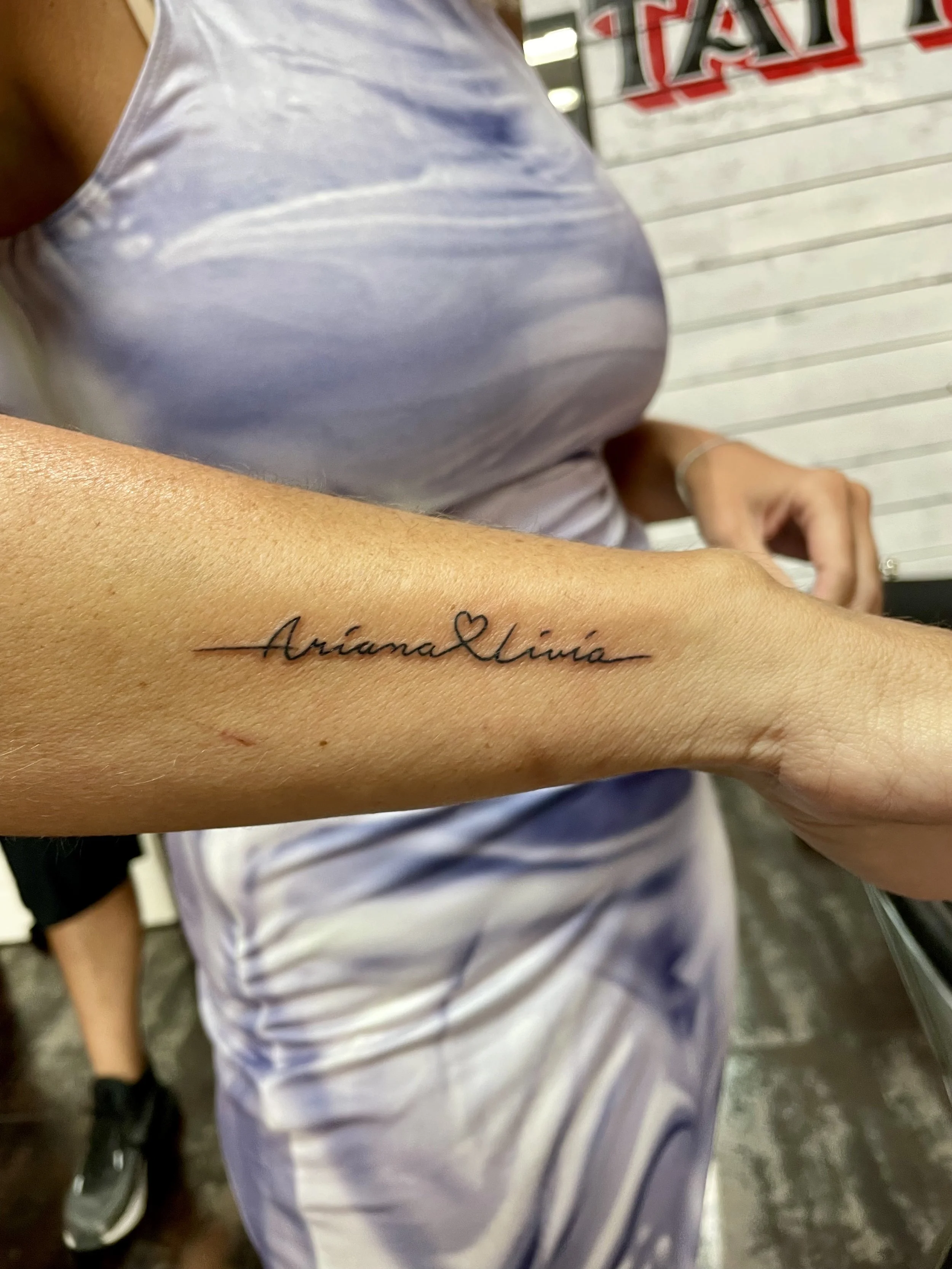 Fine line forearm tattoo featuring the names "Ariana" and "Livia" in delicate cursive lettering separated by a small heart, custom tattoo by Nick at Big Kahuna Tattoo in Boca Raton, FL.