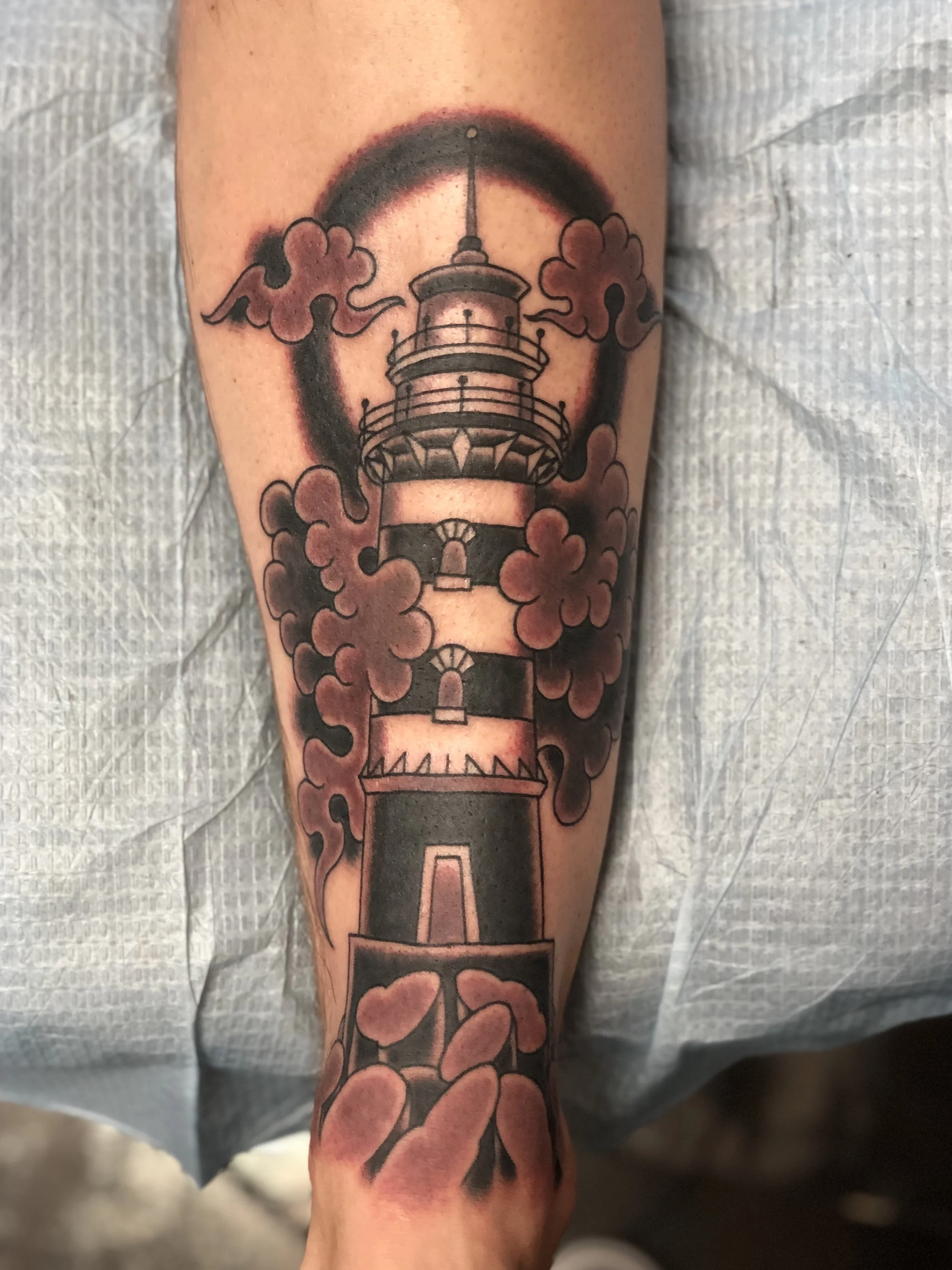 Black and grey traditional shin tattoo featuring a striped lighthouse surrounded by billowing dark clouds, custom tattoo by Nick at Big Kahuna Tattoo in Boca Raton, FL.