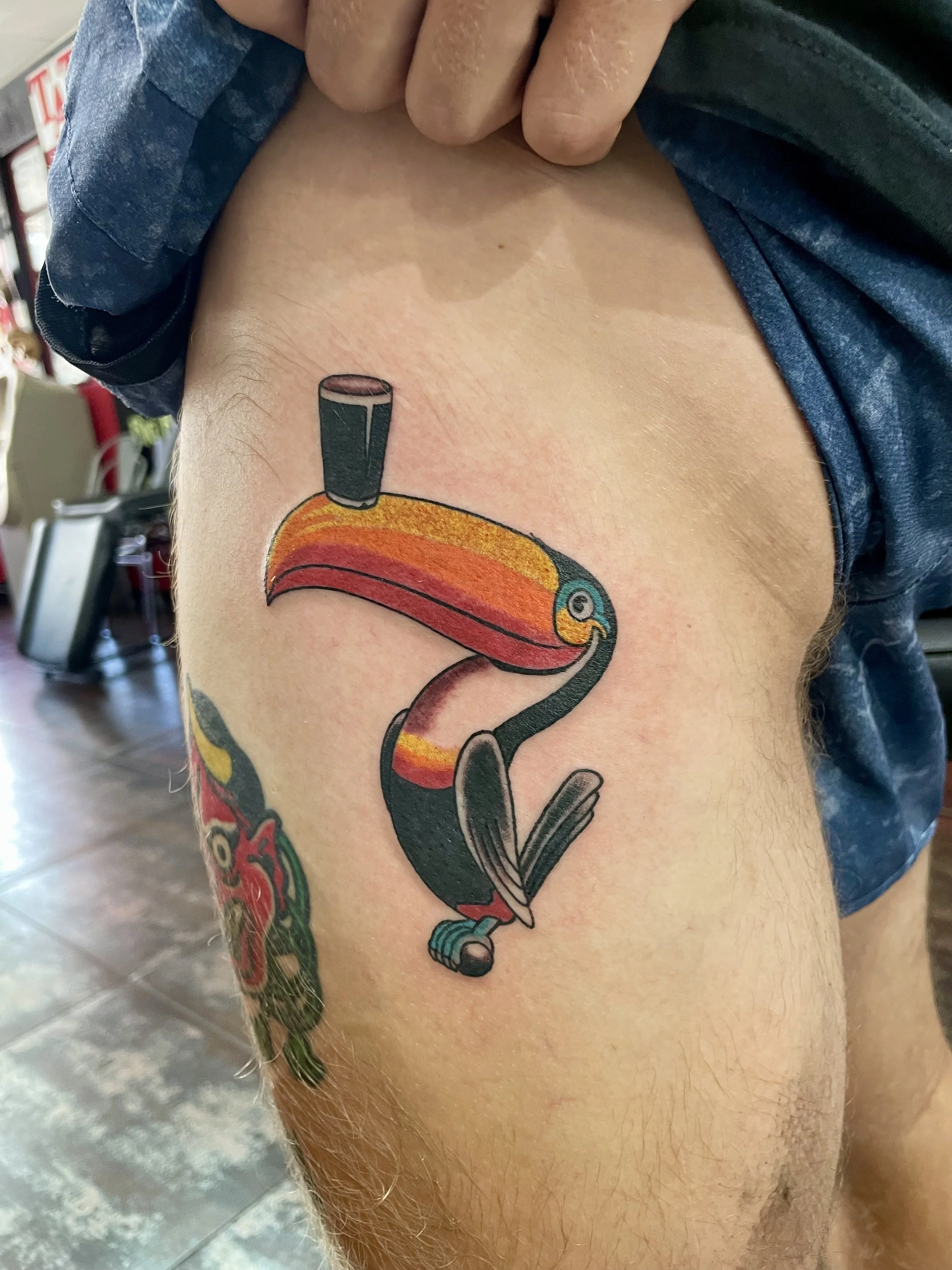 Traditional American color thigh tattoo featuring the iconic Guinness toucan with a vibrant orange and red beak balancing a pint of dark stout on top, custom tattoo by Nick at Big Kahuna Tattoo in Boca Raton, FL.