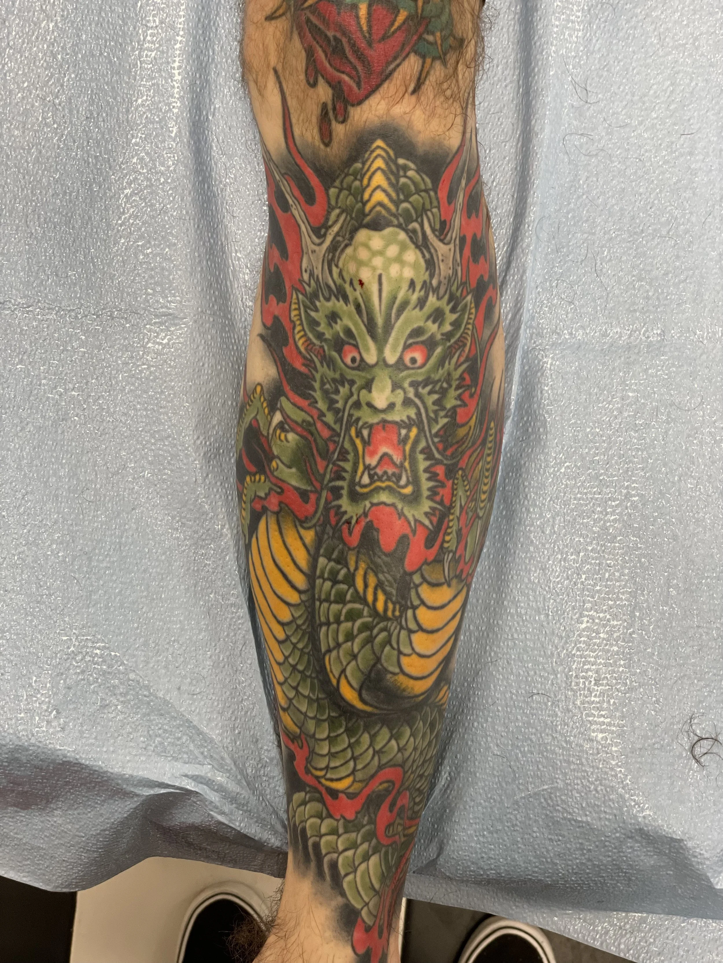 A full shin sleeve dominated by a massive green dragon with red eyes and a wide open roaring mouth, its scaled body coiling from ankle to knee surrounded by bold red flames. The kind of tattoo that takes commitment. Japanese color shin tattoo by Nick