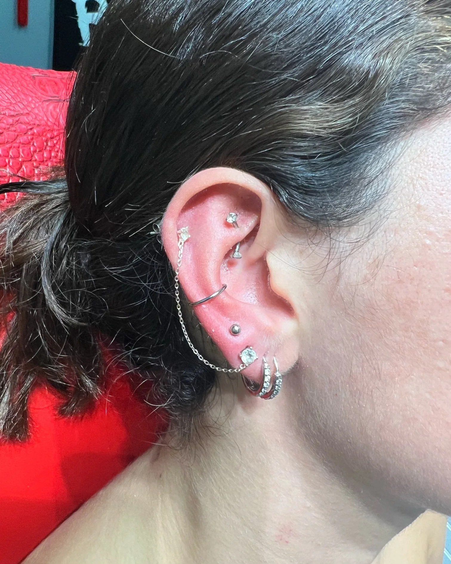 Curated ear with multiple piercings including helix, rook, tragus, and lobe placements. Featuring a silver chain connector linking upper and lower piercings, crystal studs, and decorative hoop earrings. Done at Big Kahuna Tattoo in Boca Raton.