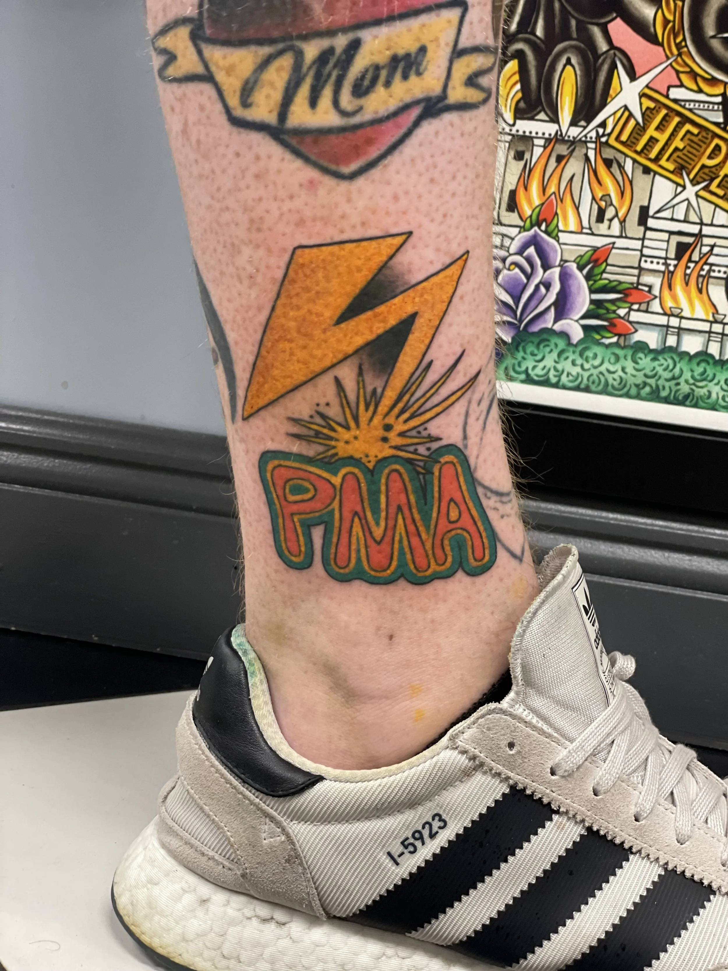 Traditional American color ankle tattoo featuring the Bad Brains-inspired orange lightning bolt with a starburst explosion above bold red lettering reading "PMA" (Positive Mental Attitude) in a green outlined bubble font, custom tattoo by Nick at Big