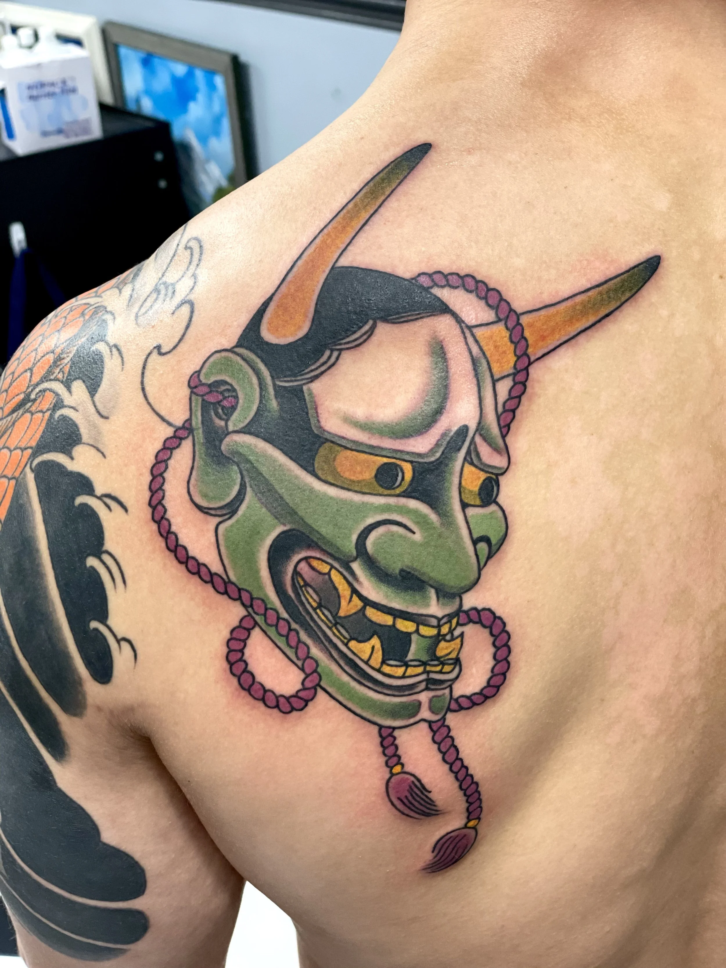 Japanese color shoulder blade tattoo featuring a traditional Hannya mask in green with golden horns, golden teeth, and dark hollow eyes, adorned with a looping purple rope and tassel, custom tattoo by Nick at Big Kahuna Tattoo in Boca Raton, FL