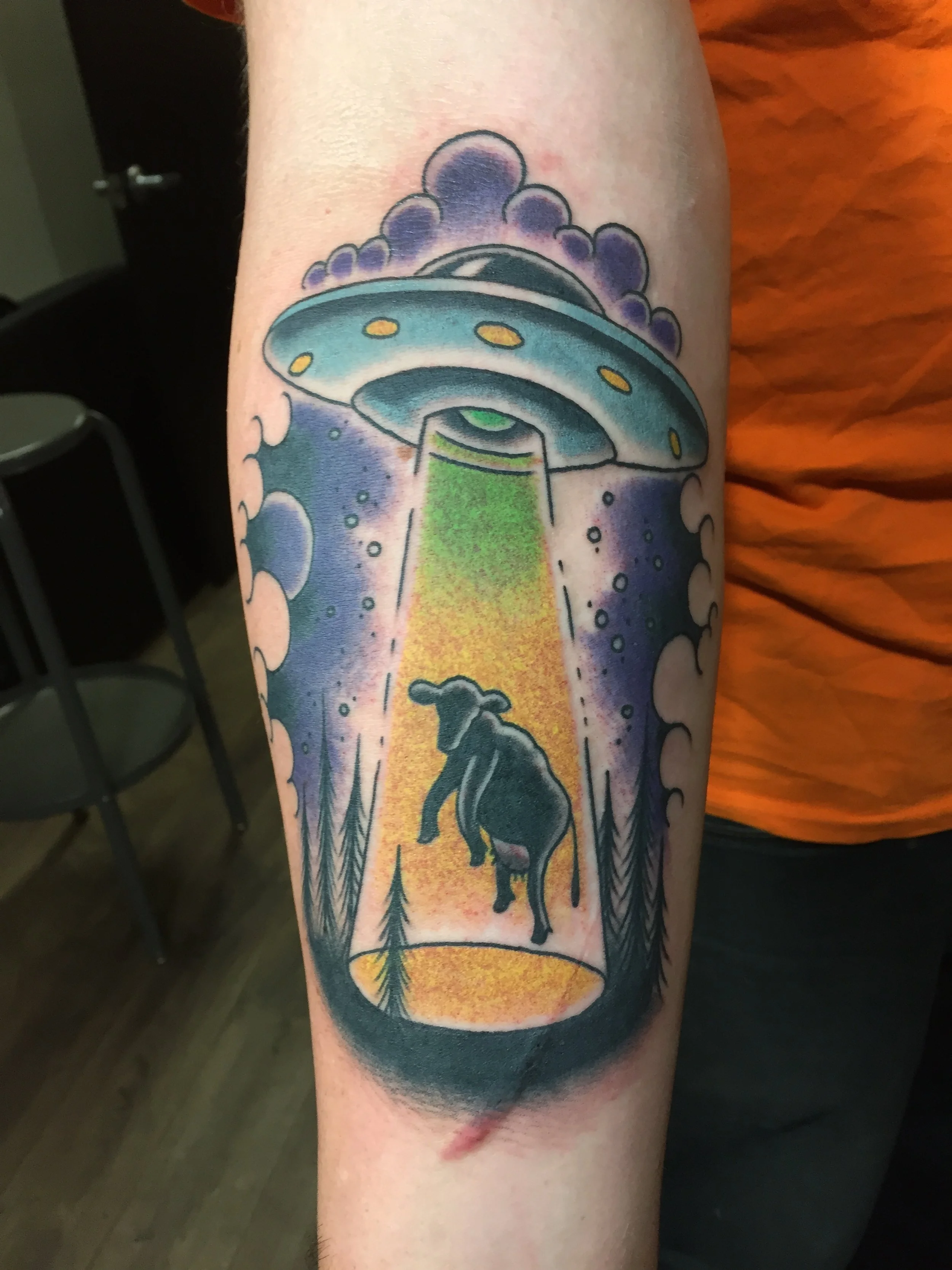 Fun and colorful traditional American forearm tattoo featuring a flying saucer abducting a cow in a green tractor beam, surrounded by purple clouds and pine trees, custom tattoo by Nick at Big Kahuna Tattoo in Boca Raton, FL.