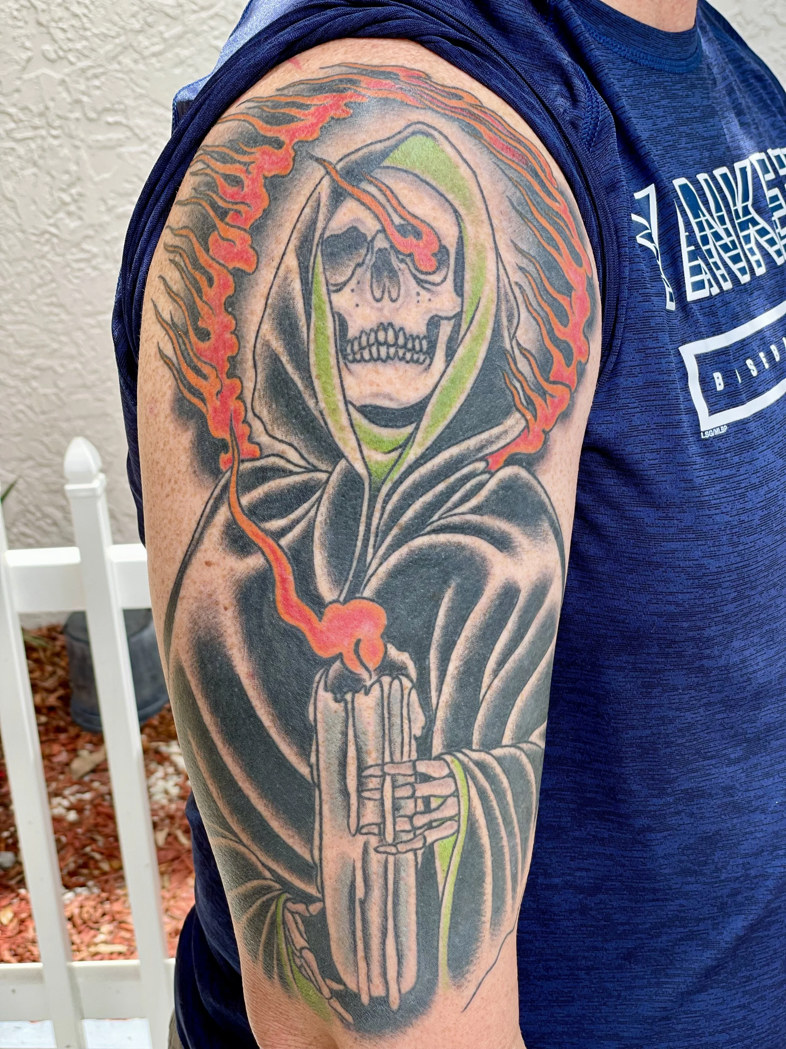 A robed Grim Reaper clutching a lit candle with skeletal hands, red and orange flames in a ring of fire, with a green trim lining the cloak. Somber subject matter drawn with the bold confidence that makes traditional tattooing last a lifetime. Color 
