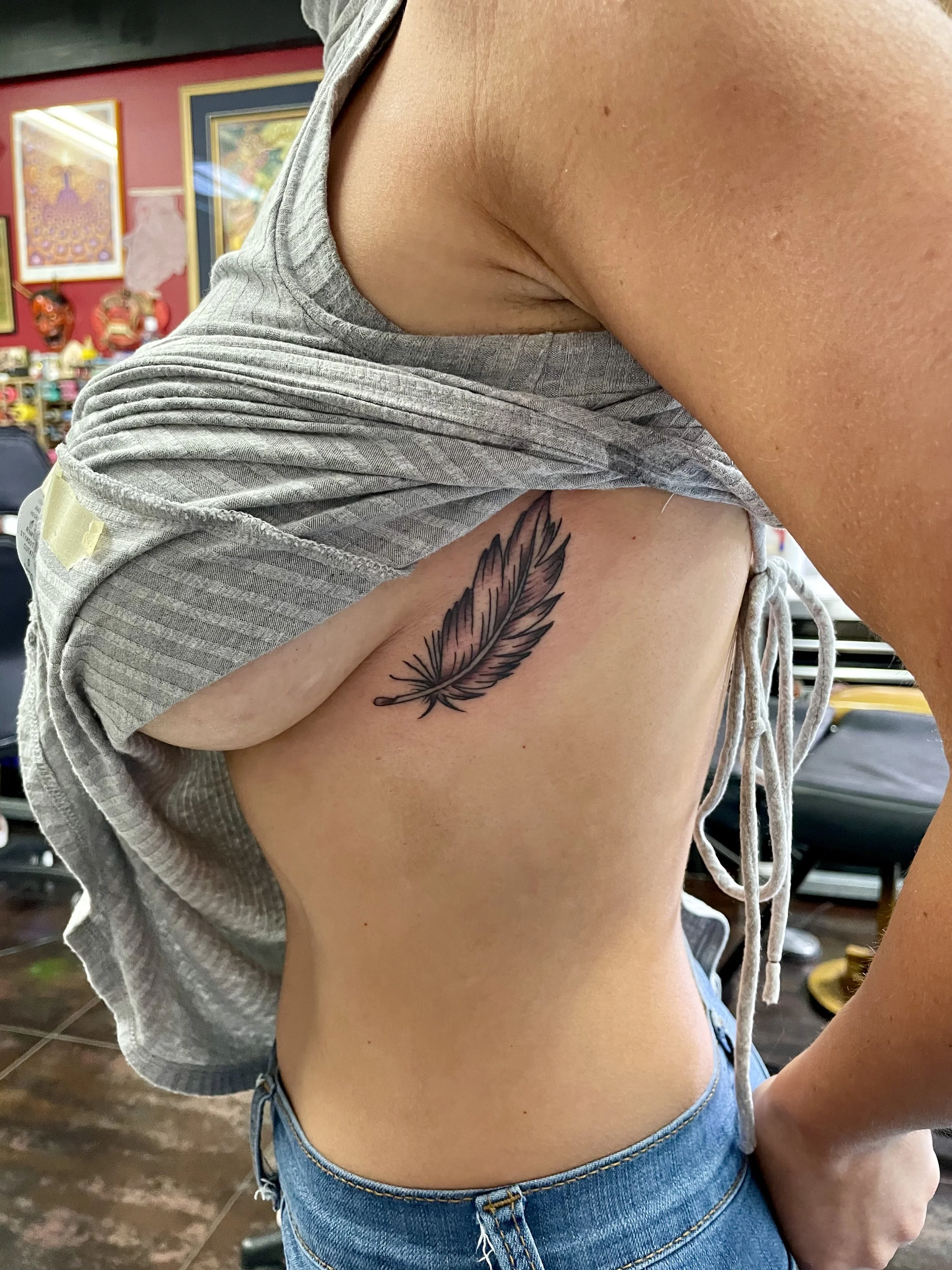 Fine line black and grey ribcage tattoo featuring a single detailed feather with soft delicate barbs and a tapered quill, custom tattoo by Nick at Big Kahuna Tattoo in Boca Raton, FL.