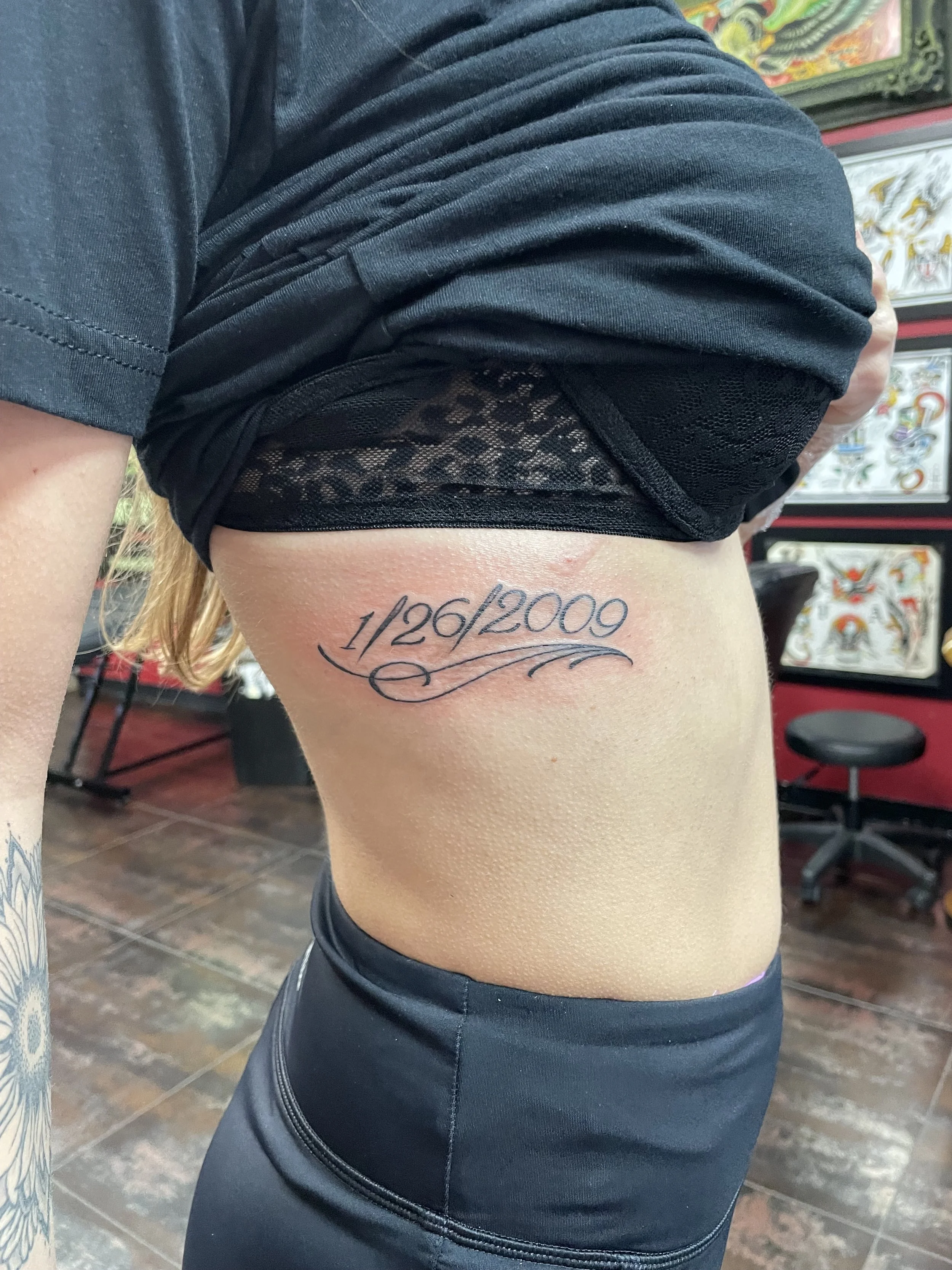 Fine line rib tattoo featuring the memorial date "1/26/2009" in elegant serif lettering with decorative flourish scrollwork flowing beneath, custom tattoo by Nick at Big Kahuna Tattoo in Boca Raton, FL.
