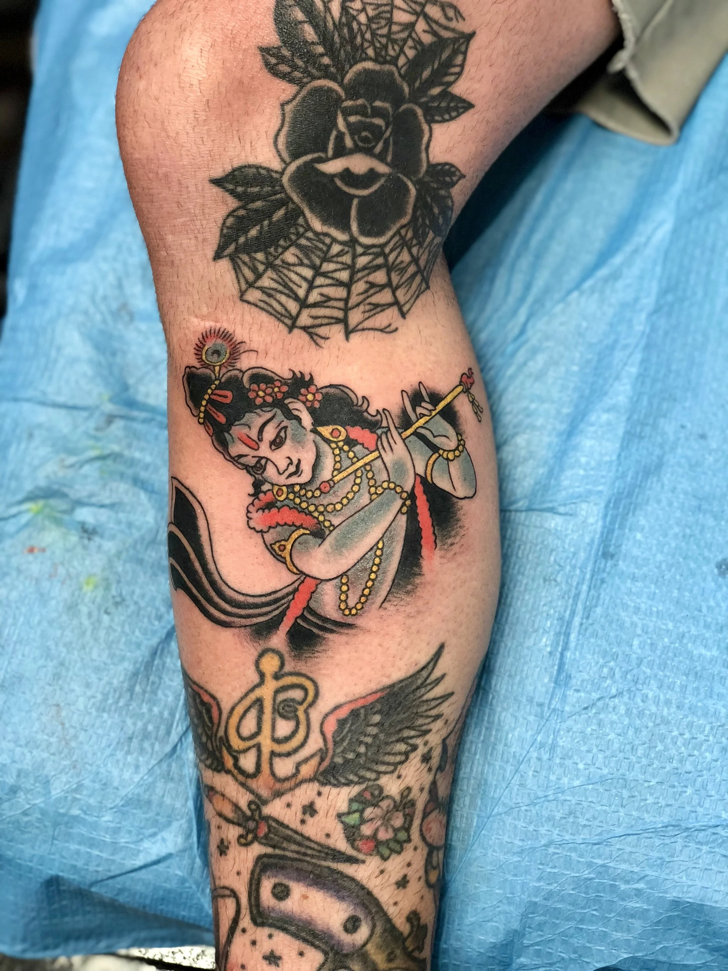 Traditional American color calf tattoo featuring the Hindu deity Krishna playing a flute with ornate jewelry and floral details, custom tattoo by Nick at Big Kahuna Tattoo in Boca Raton, FL.